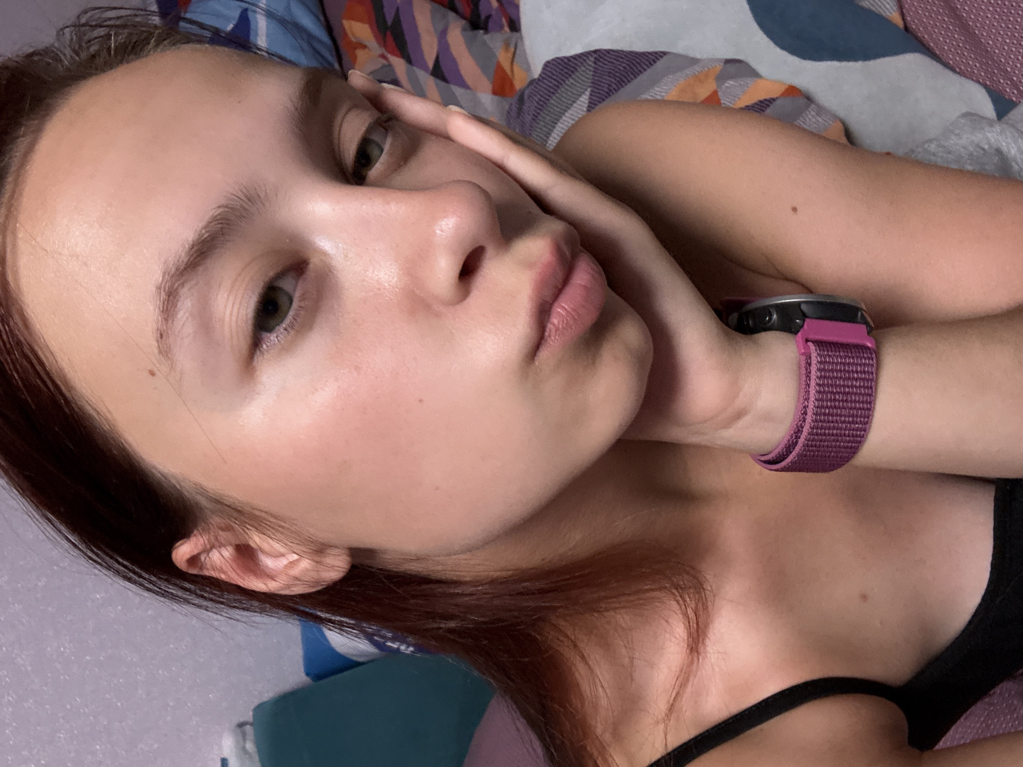 A young woman with reddish-brown hair is posing for a selfie, resting her cheek on her hand. She is wearing a black tank top and a purple watch on her left wrist. The background appears to be an indoor setting with a patterned blanket or bedding. The lighting is soft, highlighting her facial features and skin. The image was likely taken in a residential setting.