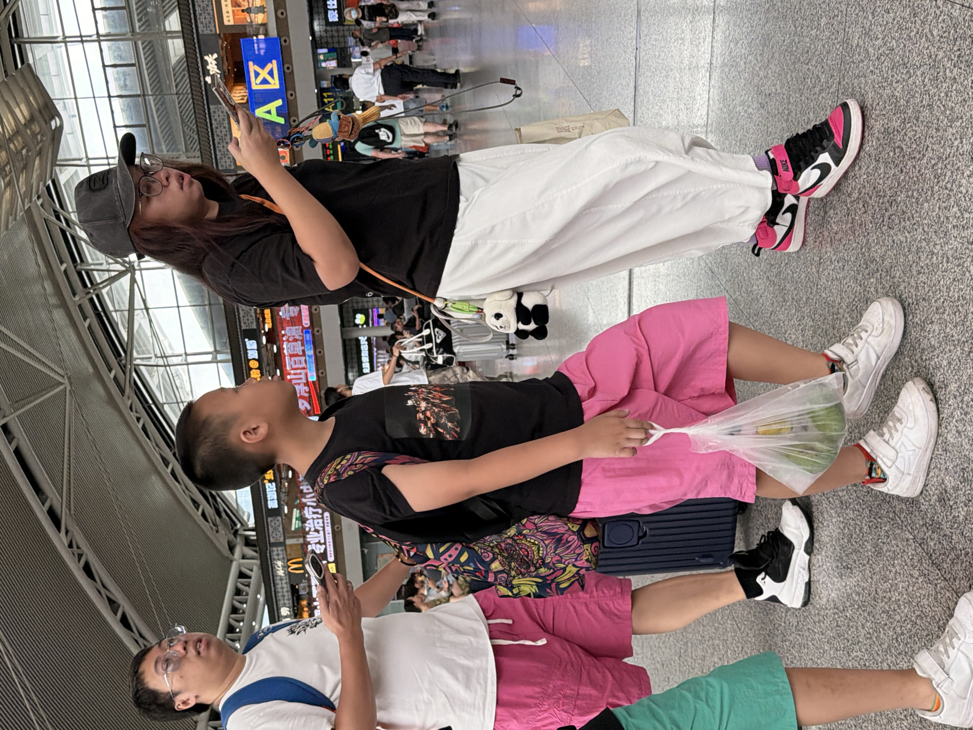 The image depicts a busy indoor environment, likely a train station or transportation hub in Panyu District, China. In the foreground, three individuals are walking. On the left, a man wearing a white t-shirt, glasses, and blue backpack is holding a phone. He is wearing bright pink shorts. Next to him, a young boy in a black t-shirt and bright pink shorts walks with a backpack and holds a plastic bag containing green fruit. To the right, a person wearing a black t-shirt, glasses, and a dark baseball cap is looking at their phone and holding a bag. They are wearing loose white pants and bright pink and black sneakers. Other individuals and luggage are visible in the background, along with signage and a high ceiling with exposed structural elements.