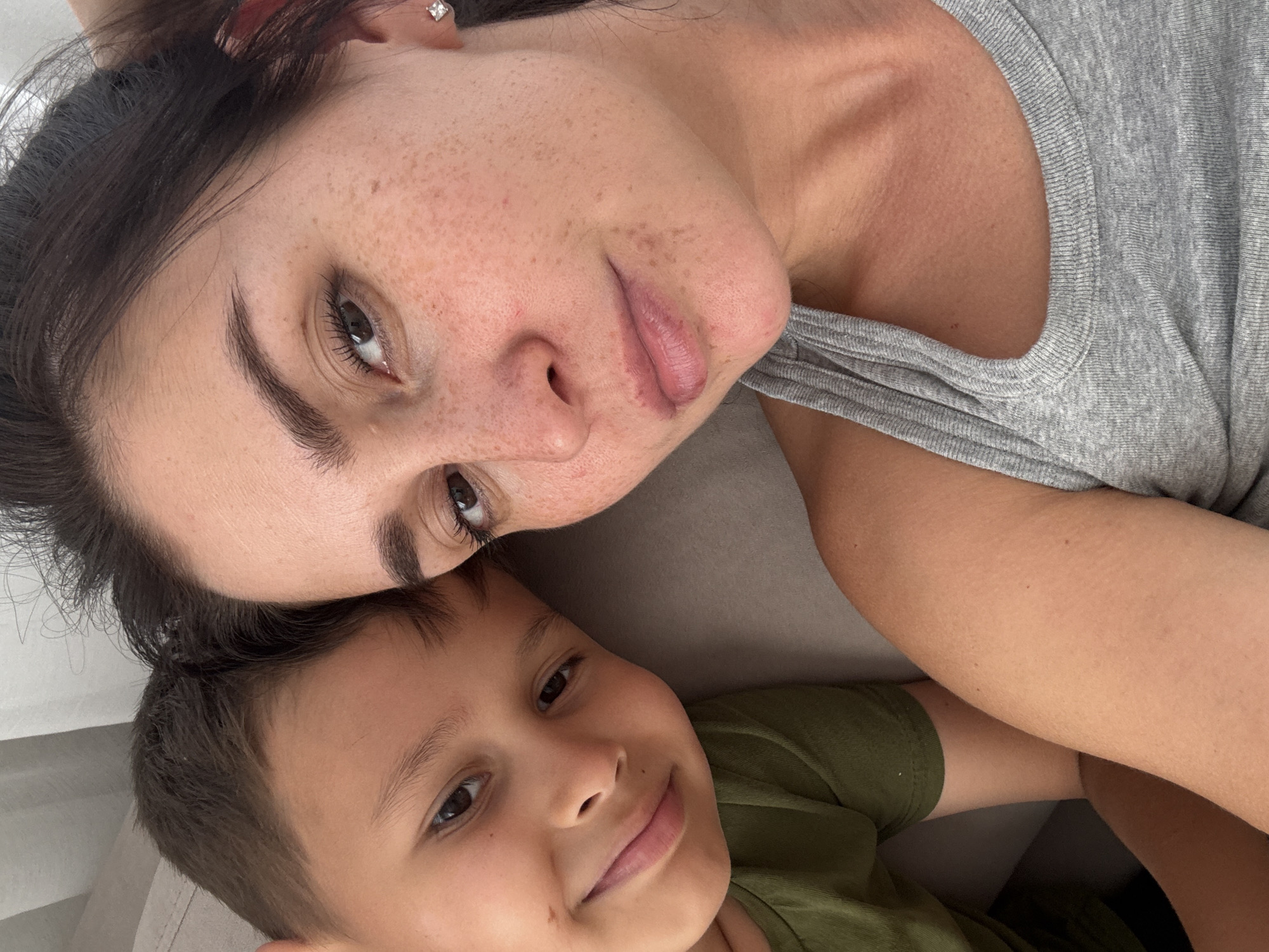 A selfie shows a woman and a young boy leaning against each other. The woman has dark hair and fair skin with freckles, and is wearing a grey tank top. The boy has brown hair and is wearing a green t-shirt. They are both looking at the camera. The background is out of focus and appears to be indoors, with a hint of a white curtain or wall. There is no indication of the location being Нижнеяркеево, Russia.