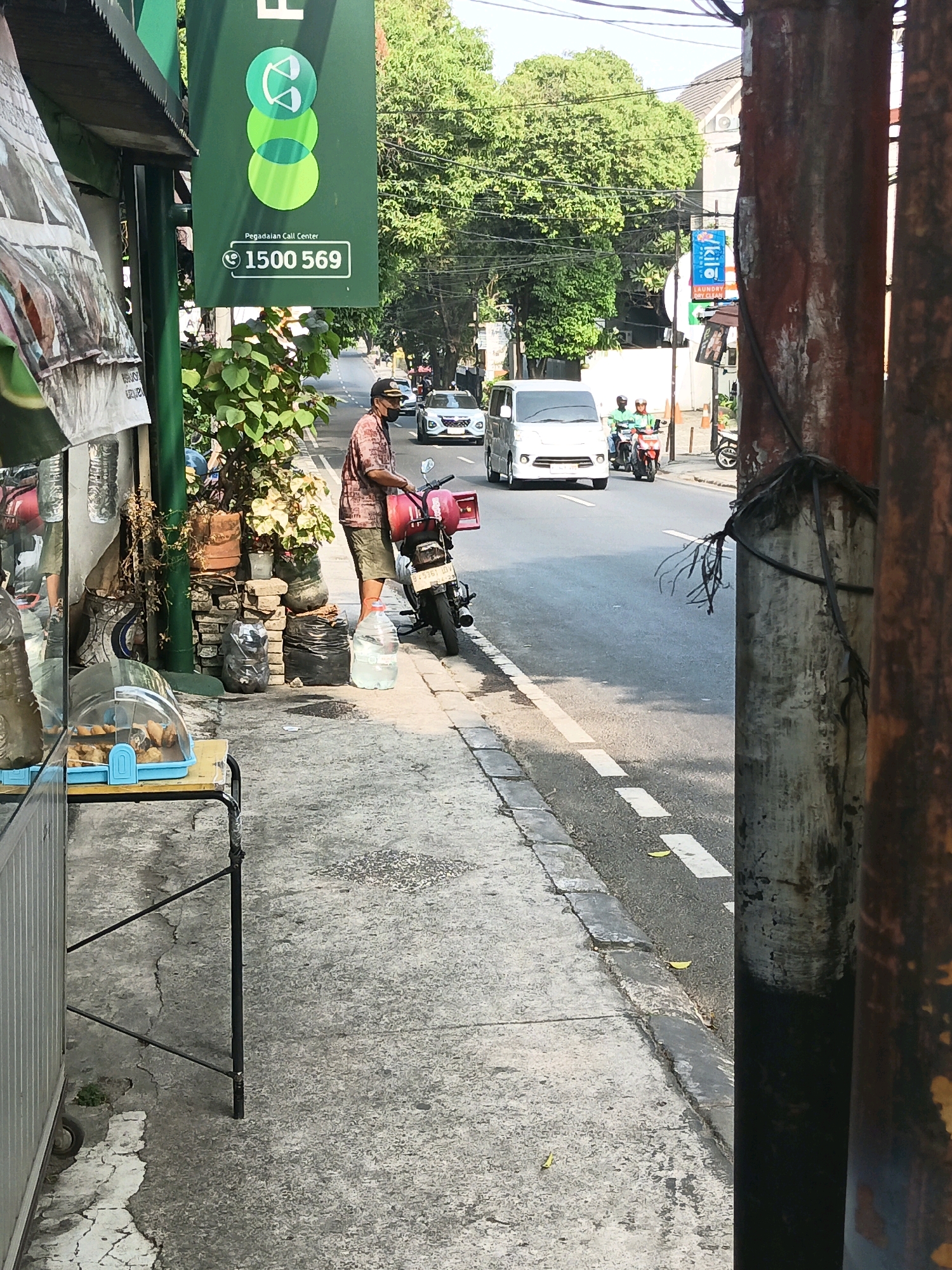 A man wearing a mask and a patterned shirt stands next to a motorcycle on a sidewalk in South Jakarta, Indonesia. He is holding a red box and a large water jug. The street behind him has traffic, including cars and motorcycles. A green sign with the text "Pegadaian Call Center" and the number "1500 569" is visible. The scene appears to be in a busy urban area with trees and buildings lining the street.