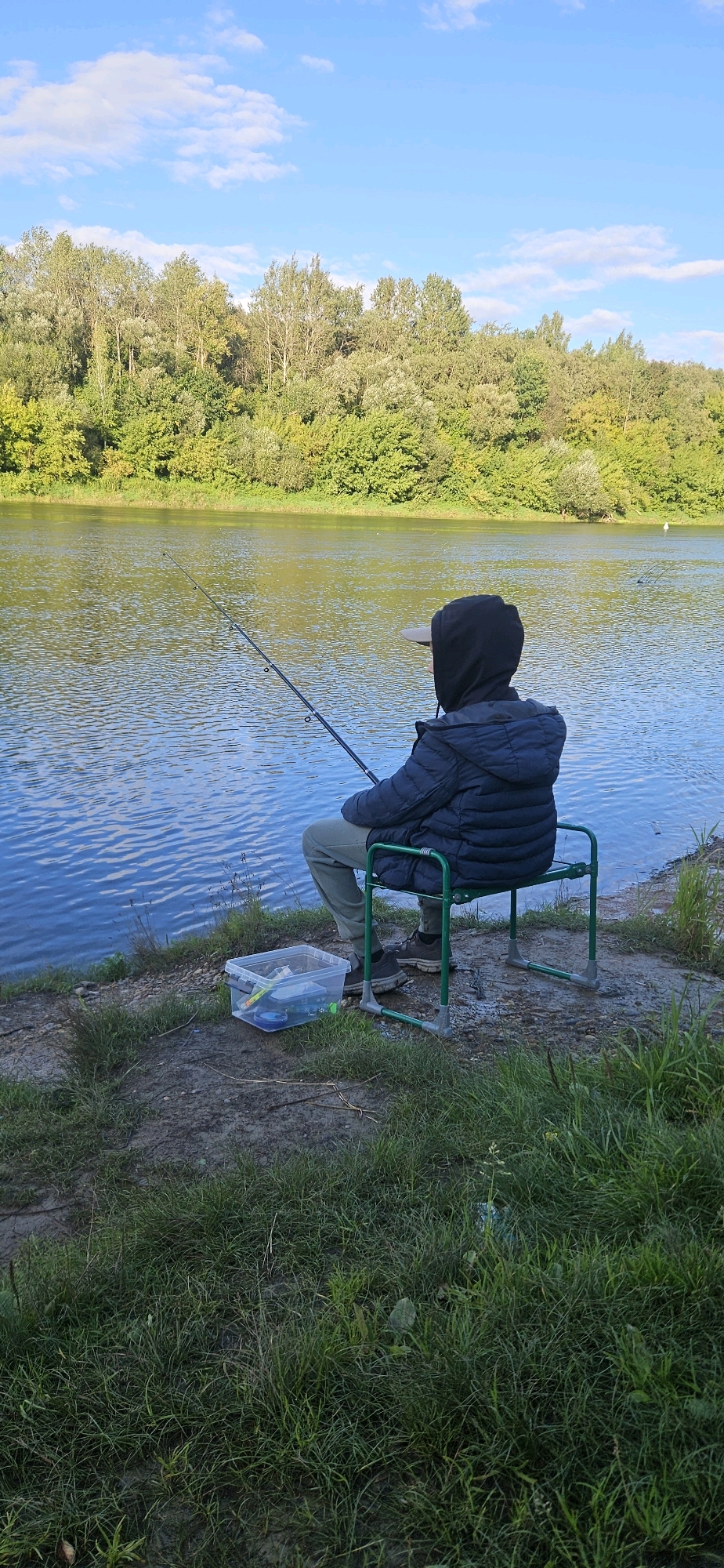 An individual, seen from behind, is fishing on the bank of a body of water. They are seated on a green folding chair, wearing a dark hooded jacket, light-colored pants, and dark shoes. A fishing rod is extended over the water, suggesting they are actively fishing. Next to the chair, on the grassy bank, is a clear plastic tackle box filled with fishing supplies. The background features a dense line of green trees and a bright blue sky with scattered white clouds. The scene is set during daylight. There is no information available to determine the specific location within Vitsebsk, Belarus.
