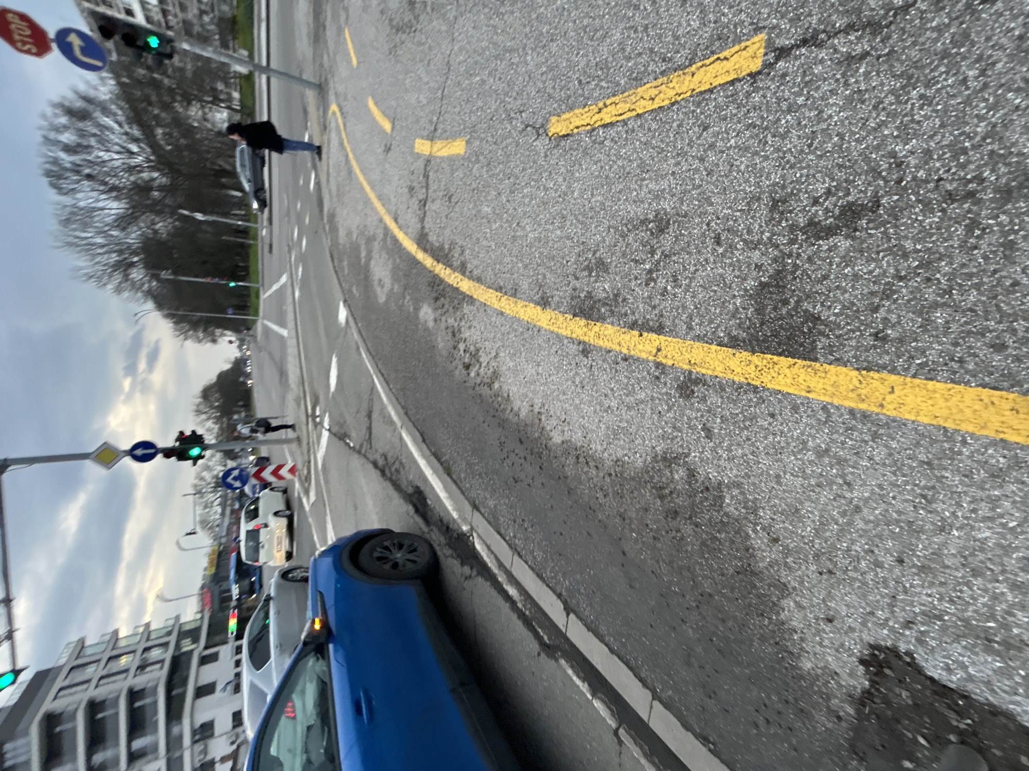 The image captures a street scene, likely in Burgas, Bulgaria, as indicated by the street signs. The viewpoint is from the edge of the road. A blue car occupies the lower portion of the frame, partially obscuring other vehicles in the background. Traffic lights are visible, showing a green light. Pedestrians are present on the sidewalks and crosswalks. Yellow lane markings divide the road. Buildings can be seen on one side of the street. The sky appears overcast.