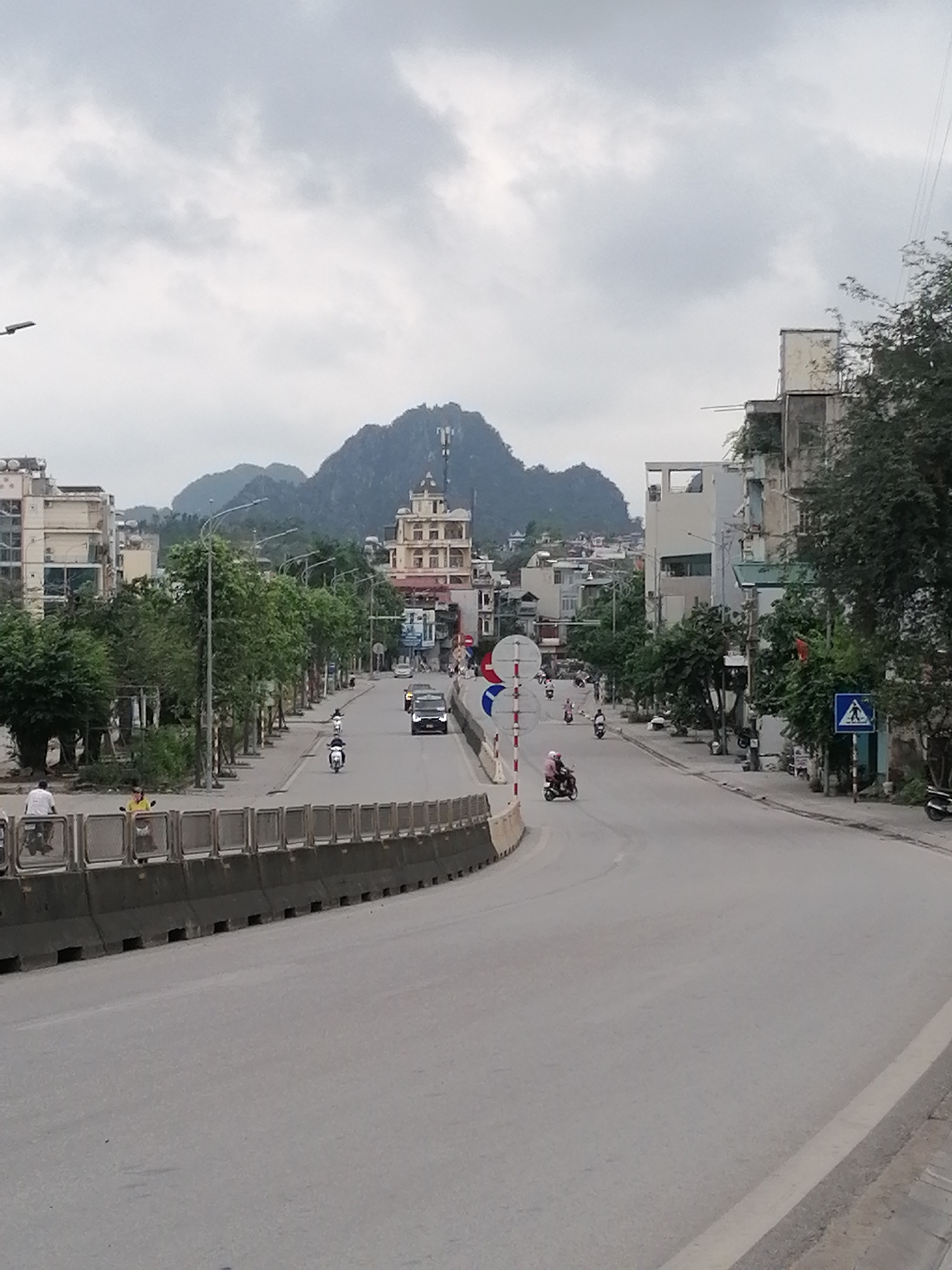 The image shows a street scene in Ha Tu Ward, Vietnam. A mountain with a telecommunications tower on top rises in the background. Buildings line both sides of a curved road, with trees interspersed. Several people are visible; some are on scooters and others are walking along the side of the road. A gray concrete barrier runs along the left side of the road. Traffic includes a black car and various scooters. The sky is overcast with gray clouds. A pedestrian crossing sign is seen on the right side of the road.