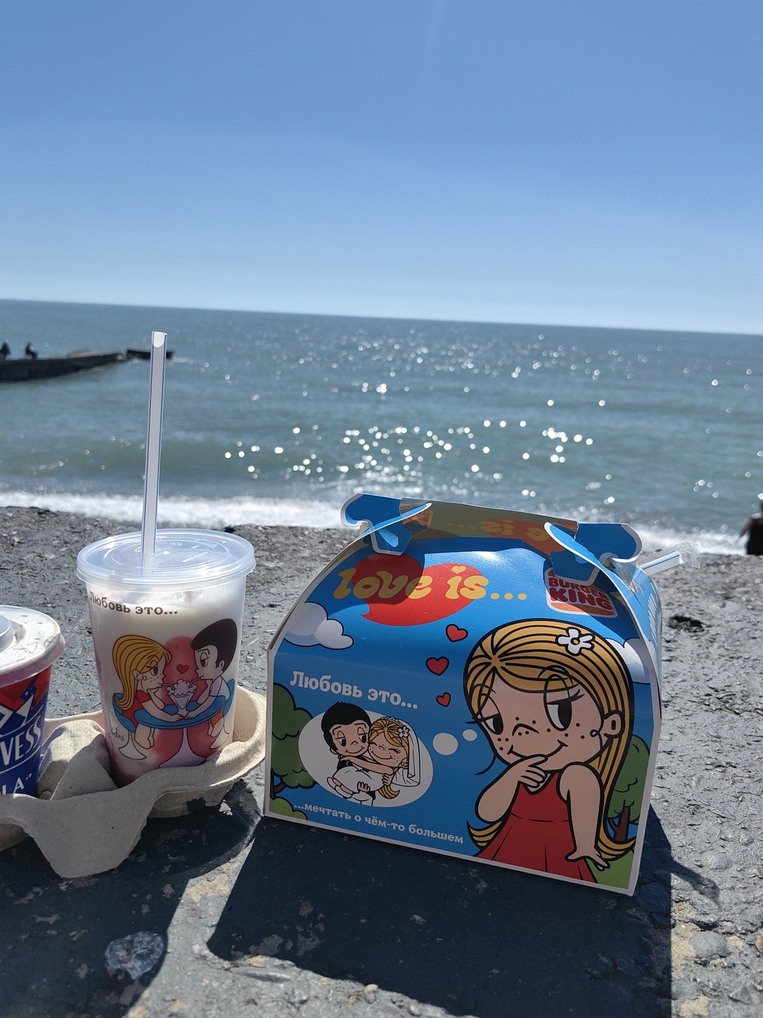 The image captures a bright, sunny day at the seaside in Sochi, Russia, showing a refreshing break by the water. In the foreground, resting on a concrete or rocky surface, are two containers for beverages. A clear plastic cup with a straw, filled with a light-colored drink like a milkshake, is prominently displayed. It features "Love is..." cartoon characters and the Russian text "Любовь это..." (Love is...). Next to it, a partially visible red and white cup shows the brand "VESS". Both cups are held in a cardboard carrier.
Beside the cups is a blue Burger King box, also adorned with "Love is..." cartoon characters. The box displays both "love is..." and "Любовь это..." along with the Burger King logo. Below the characters, the Russian text "...мечтать о чём-то большем" (...to dream of something more) is visible.
The background reveals a sparkling blue sea under a clear, bright sky. The sun reflects off the water, creating numerous shimmering spots. A pier or breakwater extends into the sea on the left, with a couple of people seated on it. The scene suggests a pleasant and relaxing moment, enjoying snacks and drinks by the water.