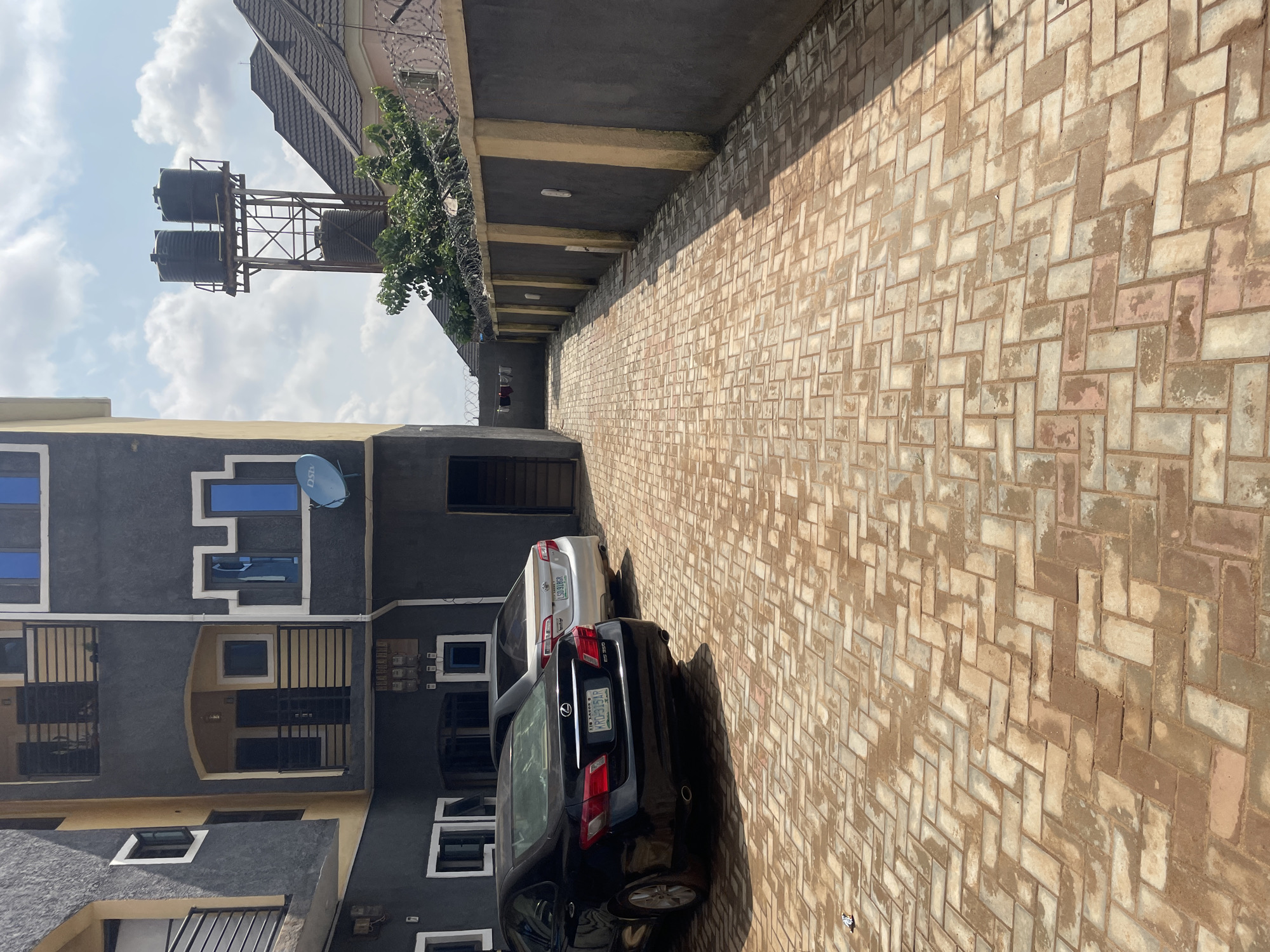 In Abuja, Nigeria, this outdoor scene depicts a paved driveway in front of a multi-story building. Two cars are parked on the patterned brick driveway: a black Lexus sedan in the foreground and a lighter-colored sedan behind it. The building, painted in dark gray and yellow, features balconies and windows. Beyond the building, a tall metal structure supports two large black water tanks, a common sight in Abuja for water storage. A concrete wall topped with barbed wire runs along the right side of the frame, separating the property from neighboring areas. The sky above is partly cloudy.
