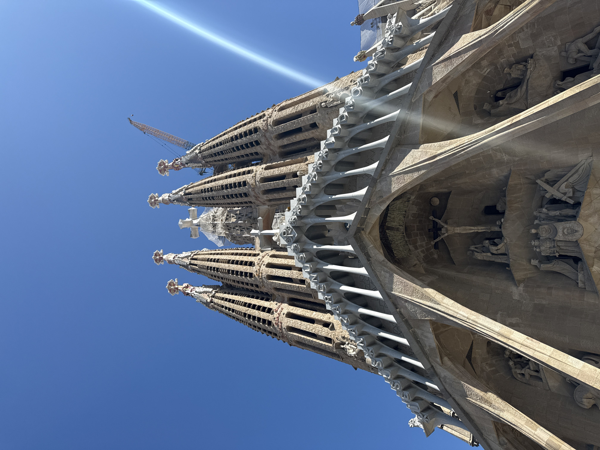 The image shows a portion of the Sagrada Familia basilica in Barcelona, Spain, angled upwards against a clear blue sky. The facade of the building is intricately detailed with carved stone. Several towers with ornate spires rise into the sky, one of which has a construction crane visible near its top. The structure features flying buttresses and numerous religious sculptures and figures carved into the stone. Light is filtering through the columns and surfaces of the architecture.