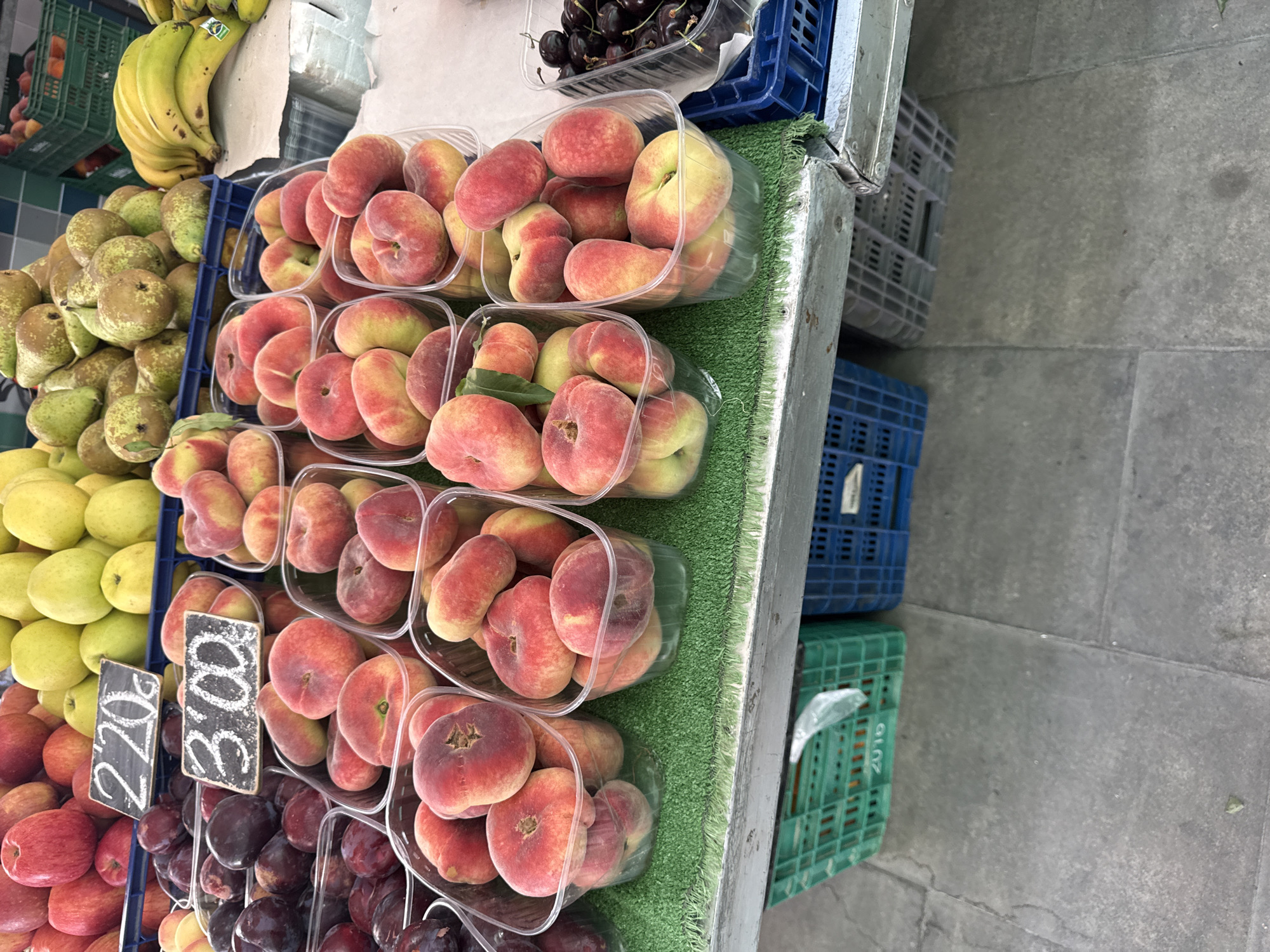 The image depicts a fruit stand with various fruits on display. In the foreground, several clear plastic containers hold flat peaches, also known as Saturn peaches. These peaches are red and yellow, with a distinct flat shape. To the left of the peaches are containers of dark red plums. Behind the peaches, there are more apples and pears, with some green apples visible. A bunch of yellow bananas is also present. The fruits are arranged on a grey surface, possibly a market stall, with green artificial turf under some of the fruit containers. Below the stall, there are several plastic crates, some green and some blue, and some grey stacked containers. The overall environment appears to be an outdoor market. There are price tags visible, one reading "2.20 €" and another "3.00 €". No people are visible in the image.