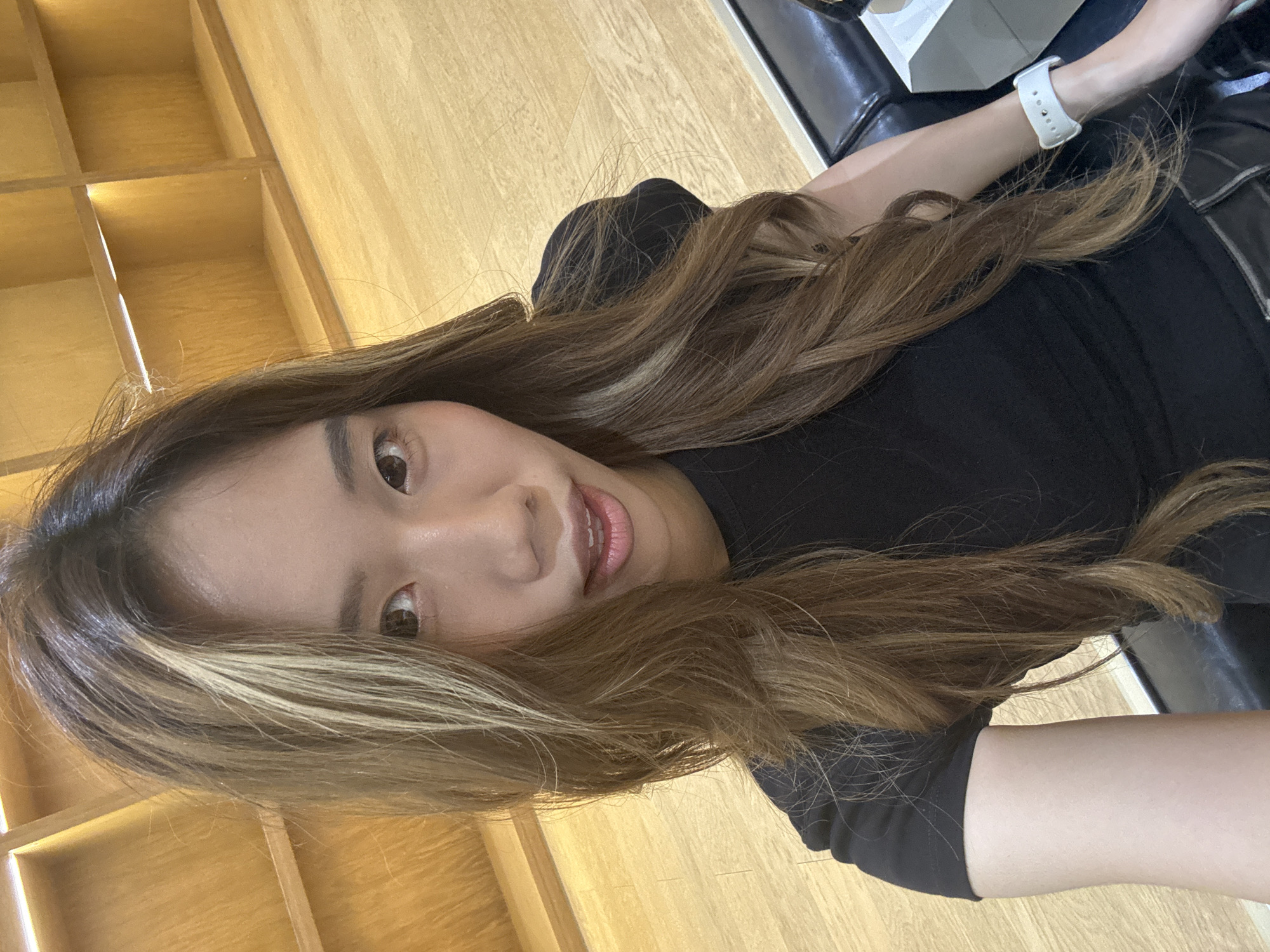 A woman with long, wavy, brown hair with blonde highlights is wearing a black shirt and a white watch on her left wrist. She is sitting in a chair that appears to be black leather, with a possible armrest visible. Behind her is a shelf made of light-colored wood with a grid pattern. The location is Nan'an District, China.