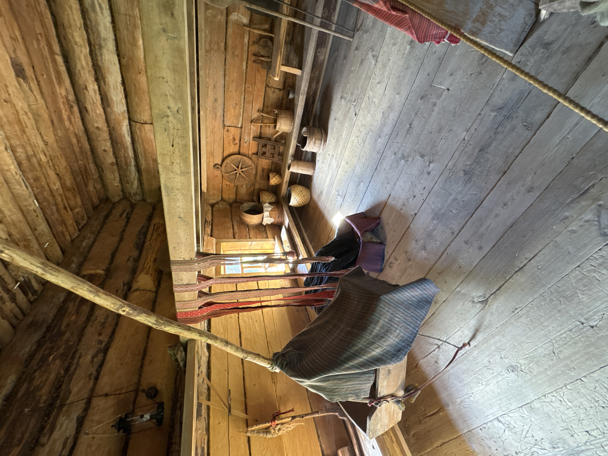 The image depicts the interior of a rustic log cabin, likely a historical exhibit, located in Gora (Khokhlovskoe S/P), Russia. The space is dimly lit, with natural light entering from a window on the right side of the frame. The walls and ceiling are constructed from rough-hewn logs, showing signs of age and wear.
In the foreground, a wooden trough-like object is positioned on the floor. A piece of striped fabric, possibly a blanket or tapestry, is draped over it, and a wooden pole extends diagonally upwards from the trough, supported by another beam. Several strips of brightly colored woven fabric, in red and beige tones, hang from a horizontal beam above the trough.
The floor is made of wide wooden planks, appearing worn and aged. Scattered on the floor are various objects, including what appears to be a rope extending diagonally across the lower right quadrant of the image.
Further into the cabin, a wooden shelf runs along the back wall, displaying an assortment of rustic items. These include woven baskets of different sizes, wooden buckets, a small wooden stool, and what looks like a primitive wooden wheel with spokes. A small ladder-like object is also present on the shelf. A lantern hangs from a beam on the left side of the image. The overall impression is one of historical preservation, showcasing traditional domestic life. No people are visible in the image.