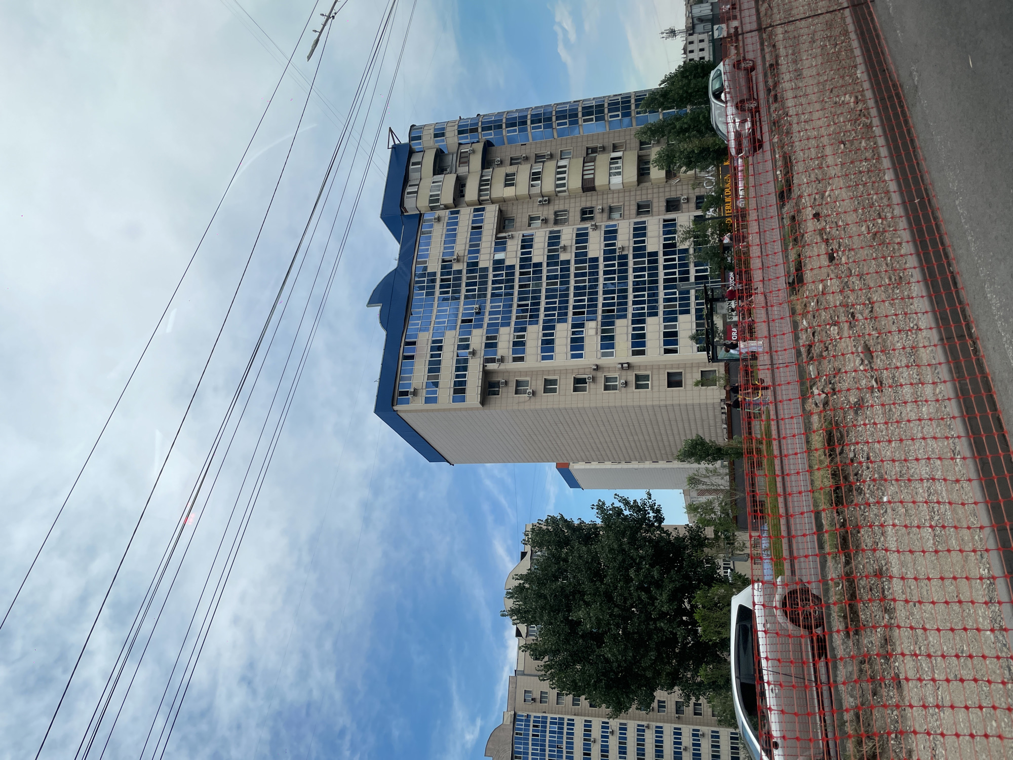 The image shows a street scene in Almaty, Kazakhstan. In the foreground, there is a red safety fence on a construction site. Beyond the fence, a silver SUV is driving on the road. In the background, there are several modern apartment buildings with blue glass windows and light-colored facades. A large green tree is visible between two of the buildings. The sky is cloudy with patches of blue. Overhead power lines cross the frame.