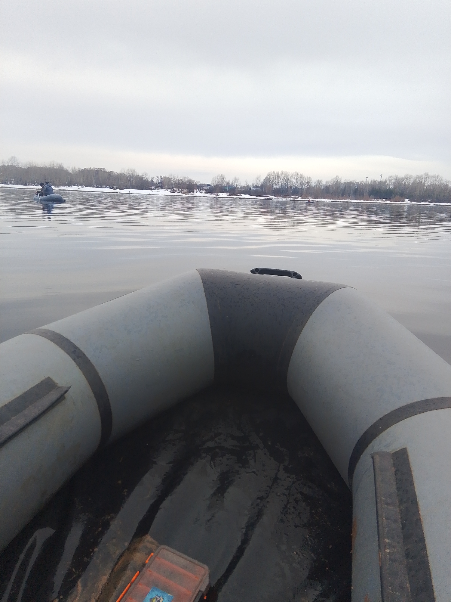 The image shows a view from inside a grey inflatable boat on a body of water, possibly a river, in what appears to be an overcast or wintery day. In the distance, there is a tree-lined bank covered in patches of snow, with some buildings visible further away. Another smaller boat with a person in it can be seen floating in the distance on the left. The foreground is dominated by the grey inflatable sides of the boat, and in the boat's floor, there's an orange and clear plastic container, likely a tackle box. Given the information provided (city of Chaikovsky, Russia), it is highly likely that the body of water is the Kama River, which is a significant geographical feature of the city.