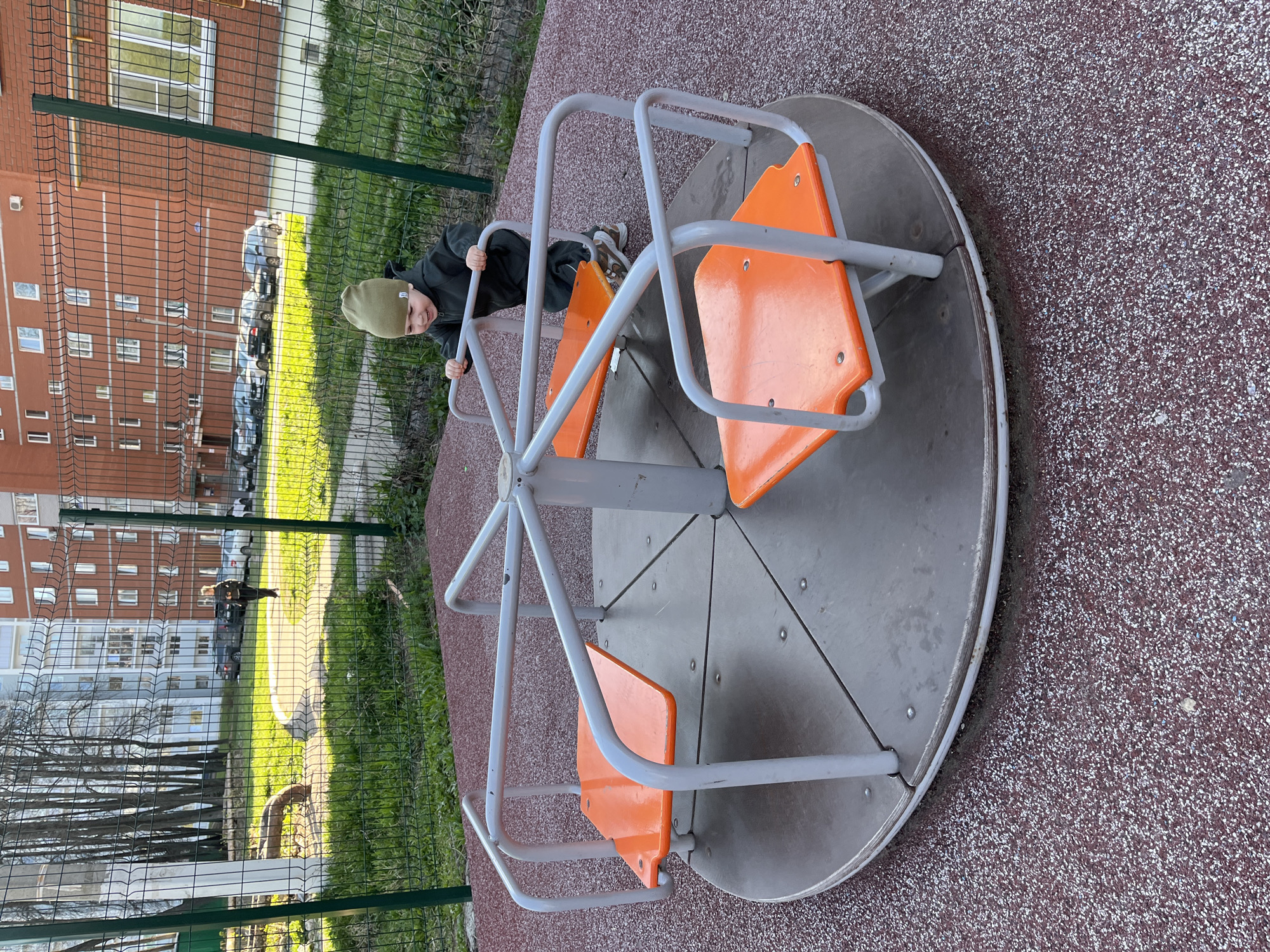 A young child, wearing a dark jacket and a green beanie, is standing on a grey and orange playground carousel. The carousel is located outdoors, surrounded by a red and brown speckled safety surface. A green fence runs parallel to a sidewalk. In the background, an apartment building is visible, along with parked cars and greenery. The location is most likely Chaikovsky, Russia.