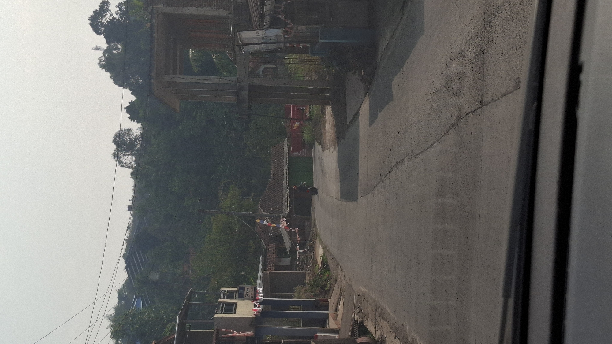 The image depicts a rural road in Cianjur, Indonesia, likely in a hilly or mountainous region, given the dense foliage covering the slopes in the background. The road is paved and appears to be a two-lane street with a slight curve. On the left side of the road, there is a building with an ATM machine and flags displayed. Further down the road, several houses with traditional Indonesian roofs are visible. On the right side, there is a partially constructed building with concrete pillars and a roof. A person is riding a motorcycle on the road, heading away from the camera. The sky is hazy, suggesting either a cloudy day or air pollution. The overall impression is of a quiet, possibly developing, community in a tropical environment.