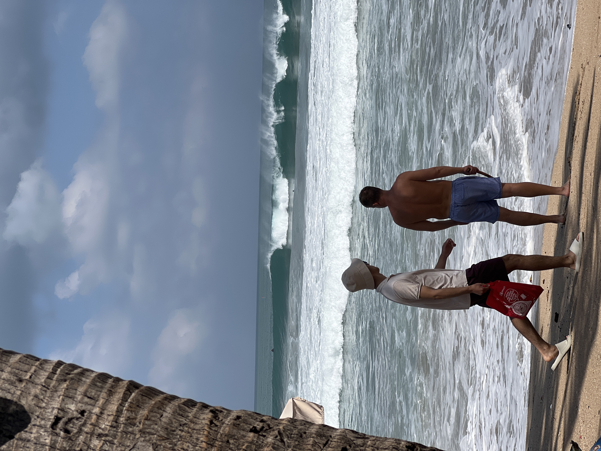 Two men stand on a sandy beach in Kuta, Indonesia, facing a powerful surf. The man on the left, wearing a light-colored collared shirt, maroon shorts, and a wide-brimmed hat, carries a red tote bag. He is walking to the left, with his left leg forward. The man on the right, shirtless and wearing blue striped shorts, faces the ocean, holding a thin stick in his right hand. He stands with his back to the viewer. The ocean exhibits large, crashing waves with white spray, suggesting a strong current and active surf. In the distance, surfers are visible in the water. The sky is cloudy, with a mix of gray and lighter patches. The foreground shows the sandy beach with shadows cast by what appears to be a palm tree on the left side of the frame.
