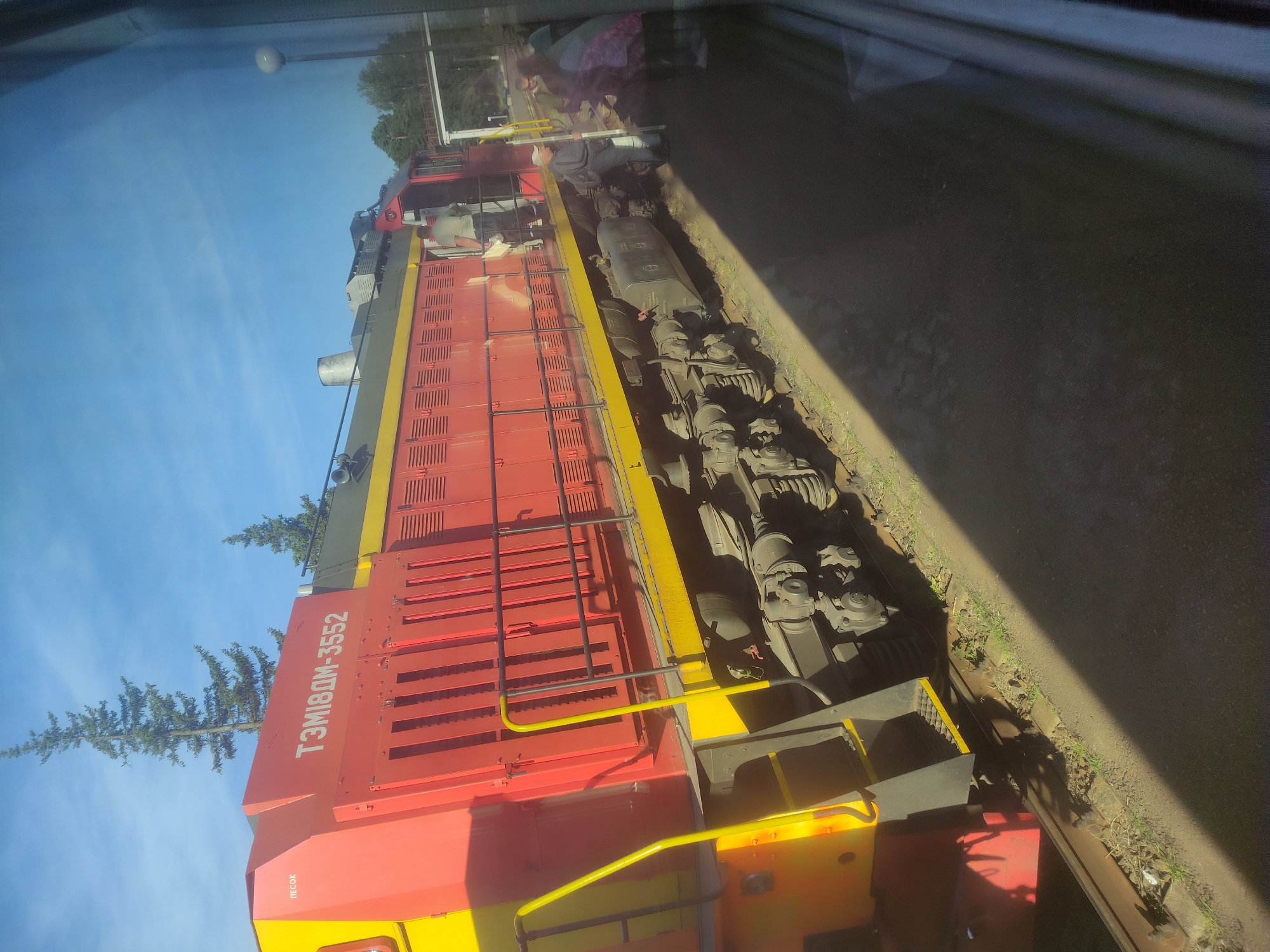 The image shows a red and yellow locomotive with the number ТЭМ18ДМ-3552 displayed on its side. The locomotive is parked at a station platform, indicated by the presence of a track and a paved area. Several people are visible on the platform near the locomotive. One person, wearing a light-colored shirt, is standing by the open door of the locomotive. Another person, wearing a cap and dark clothing, is standing on the platform holding a long, thin object. Further down the platform, a group of people, mostly blurred, are gathered, with one individual wearing a pink garment. The environment is outdoors, with a clear blue sky and trees visible in the background. The scene appears to be a sunny day. The presence of a train at a station suggests it is a transportation hub within the city of Mahilyow, Belarus.