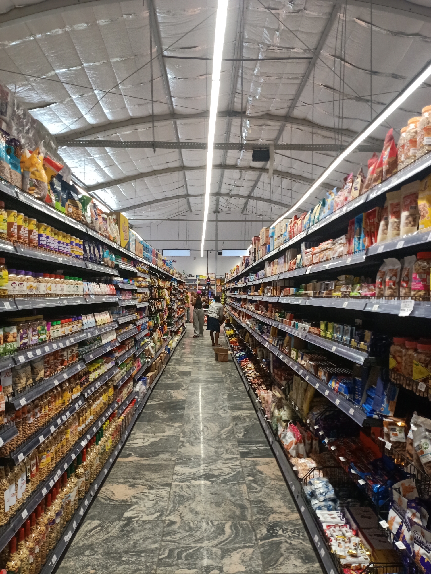 The image shows a view down a grocery aisle. Shelves are stocked with various food products including nuts, snacks, and beverages. Several people are visible farther down the aisle. The floor is a light grey with a marble pattern. The ceiling is high and made of metallic insulation panels. A long fluorescent light fixture runs down the center of the aisle. The location is a grocery store in a city of Unknown, Unknown.