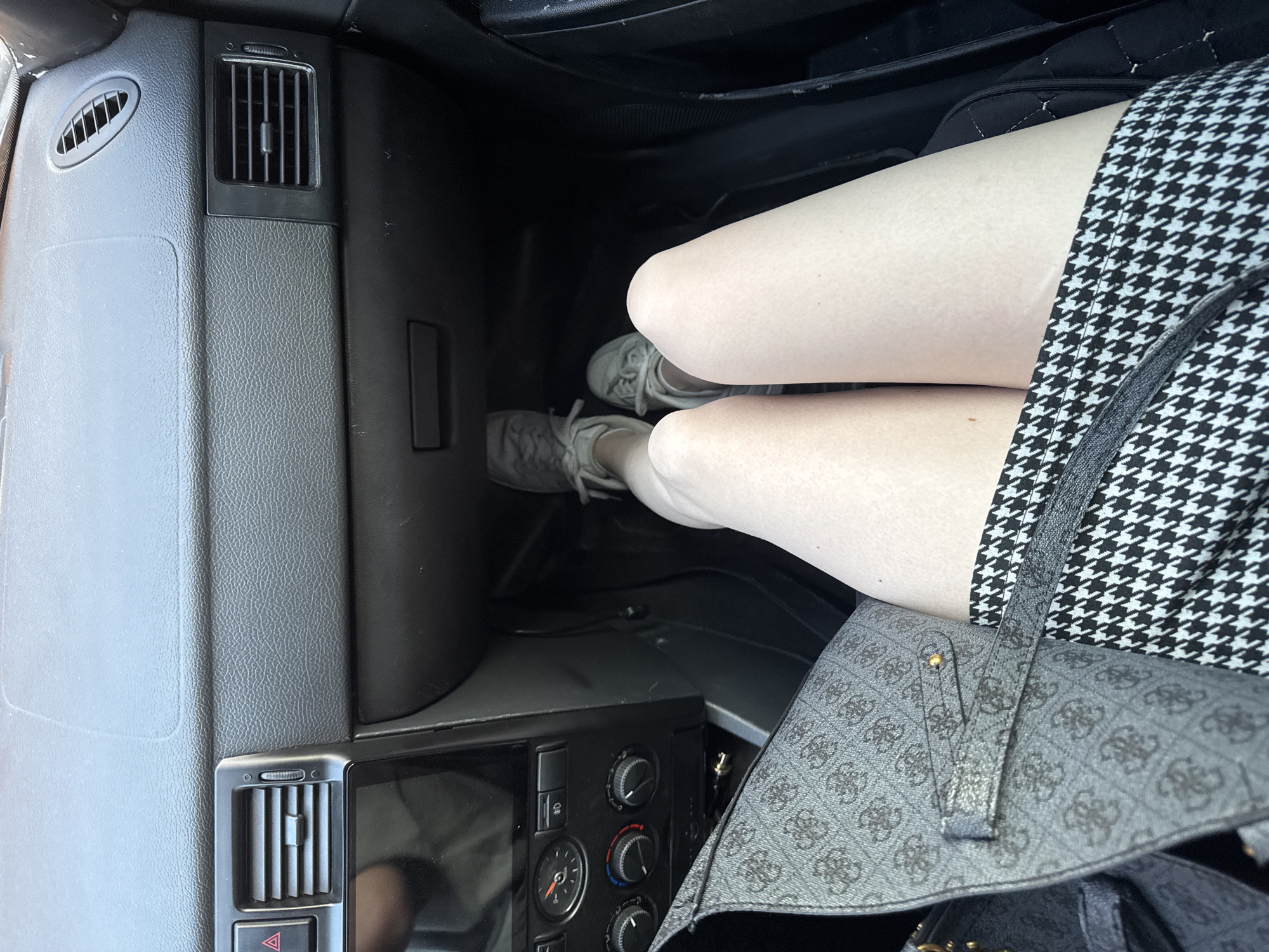 A view inside a vehicle from the passenger seat looking towards the floor. The vehicle's dashboard, including air vents and climate control panel, is visible. A person's legs, clad in light-colored tights or stockings and a black-and-white houndstooth patterned skirt, are positioned in the center of the view, with gray sneakers on their feet. A gray purse is adjacent to the legs. The location in Perm, Russia cannot be determined from this image.