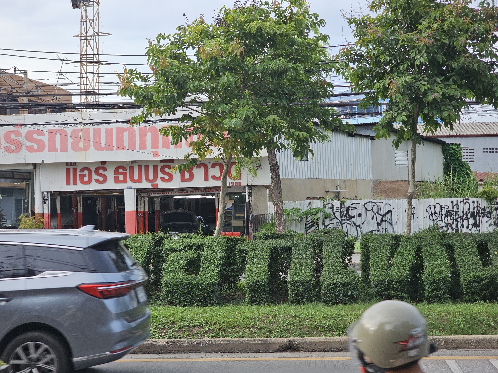 In Bangkok, Thailand, a gray SUV is captured in motion on a street. Ahead of the vehicle, a car repair shop with red and white signage is visible. Trees line the street, and a hedge sculpted into large green letters spelling out "ATTRACTION" stands prominently in front of the shop. The background features buildings, overhead power lines, and a metal fence with graffiti. A person wearing a helmet is partially visible in the foreground.