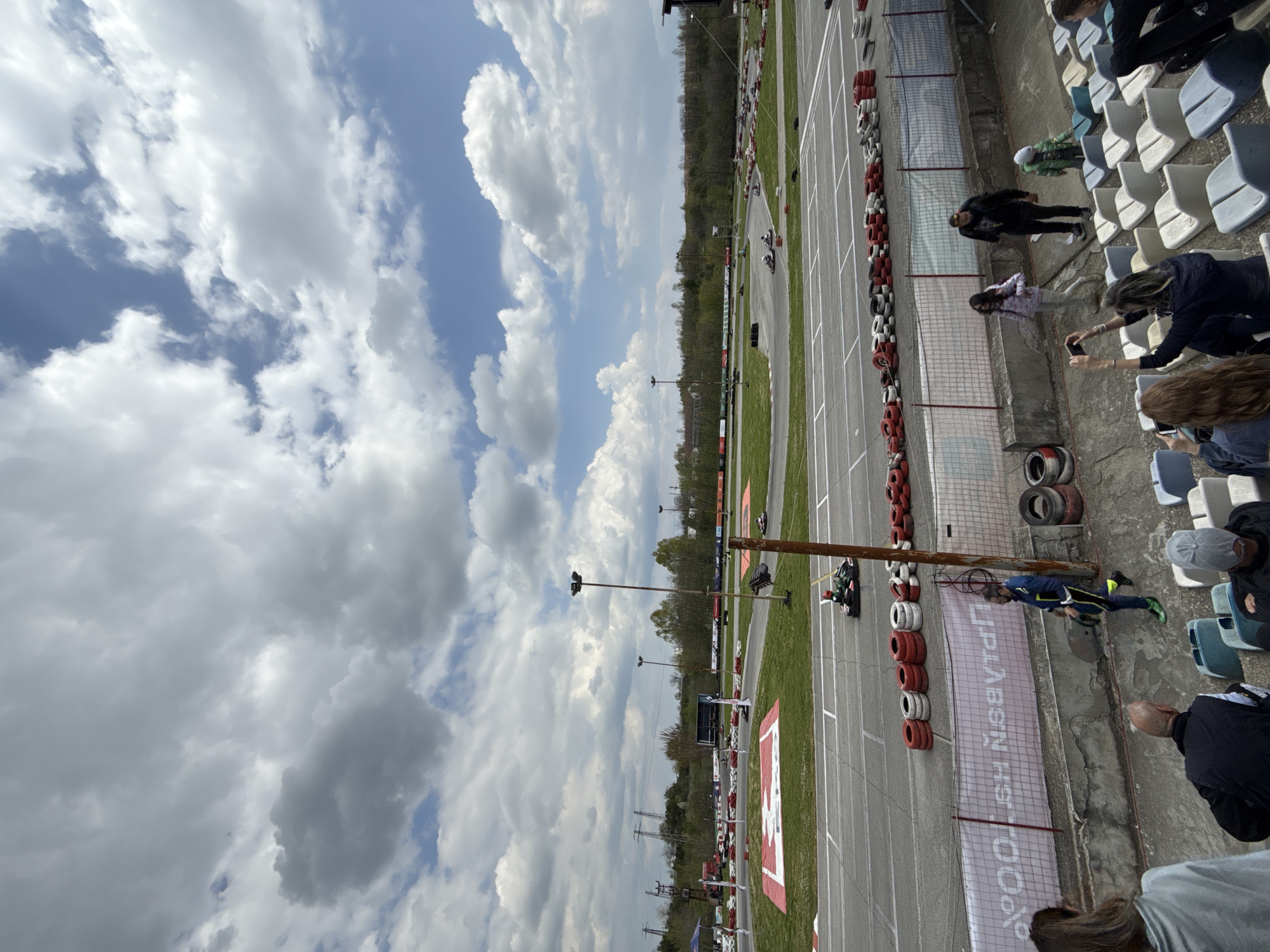 A motor racing track near Haskovo, Bulgaria, is viewed from a spectator stand. A person in racing attire lies on the ground near the track's edge, adjacent to a safety barrier. Spectators observe from the stands, with some using phones to capture the event. Karts are racing on the track. Safety tires line the track's border, and a banner is displayed along the side. The sky is partly cloudy.