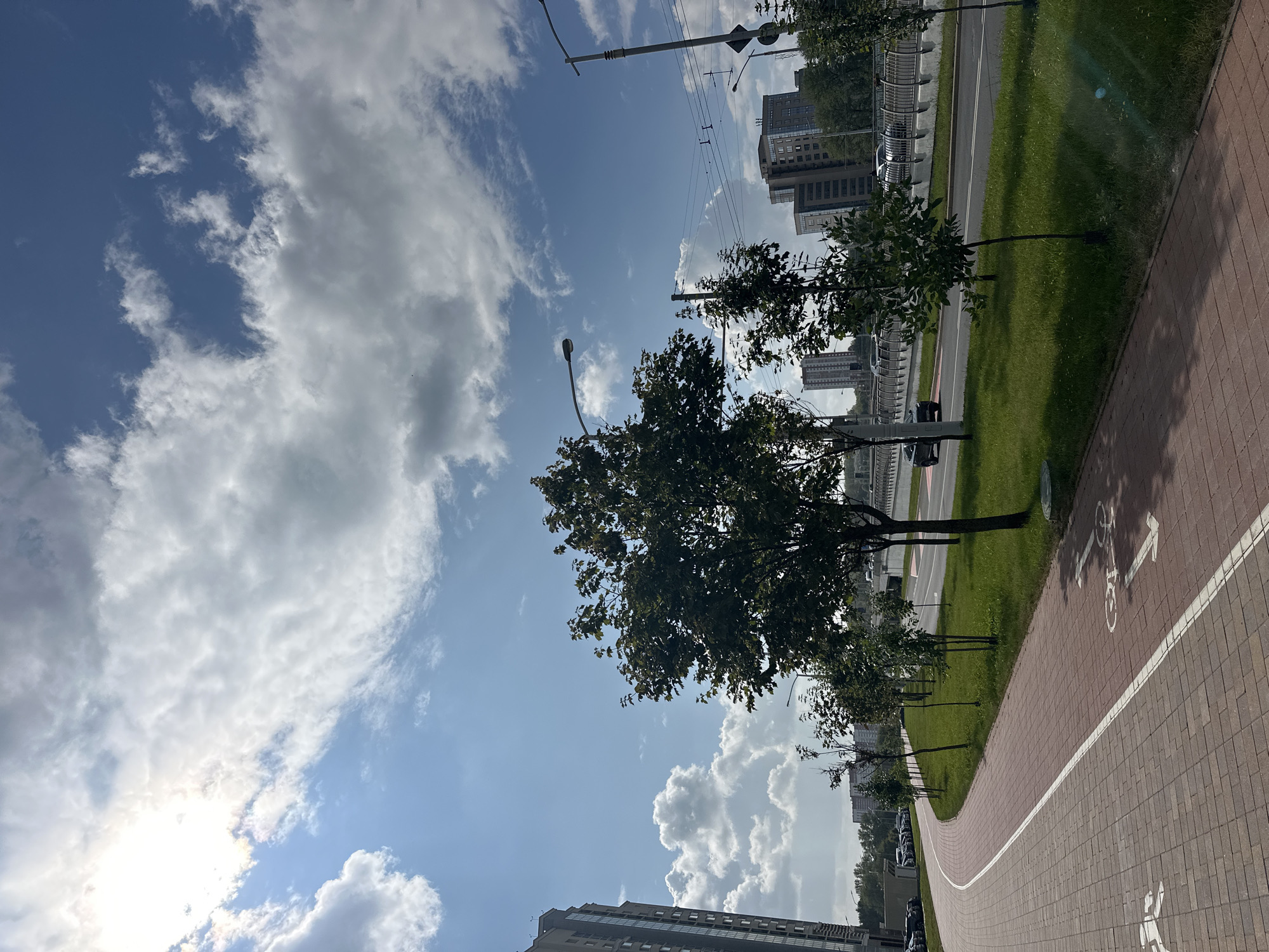 This is a landscape photograph taken on a sunny day with scattered clouds in the sky. In the foreground, there is a red brick paved bicycle path with white lane markings. The path curves to the left. To the right of the path is a grassy median strip with several young trees and a larger, mature tree. Beyond the median is a multi-lane road with traffic. In the background, there are several modern buildings, including a tall, slender building on the left and other multi-story buildings further back. Streetlights are visible along the road. The scene appears to be an urban environment in Minsk, Belarus, likely a parkway or a road with dedicated bike lanes. There are no people visible in the image.