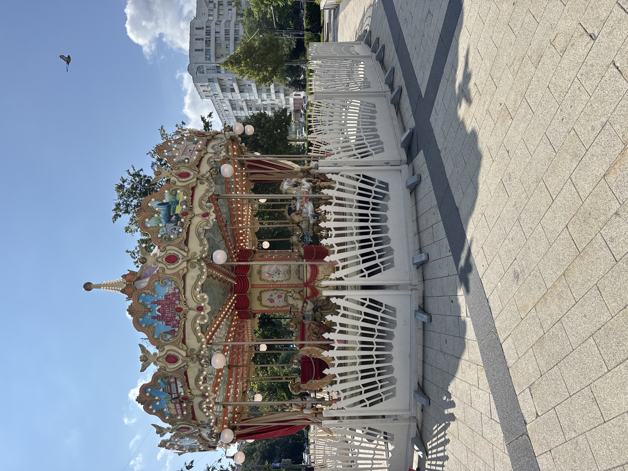 A carousel stands in a city square on a sunny day. The carousel is brightly decorated with ornate details, including painted panels depicting architectural scenes, figures of cherubs, and numerous lights. The carousel features several horses and other decorative elements. A white, decorative fence surrounds the base of the carousel. The ground is paved with light-colored square tiles, with a dark gray strip running through it. In the background, there are trees, modern apartment buildings, and a bird flying in the clear blue sky. The scene is set in Zelenograd, Russia.
