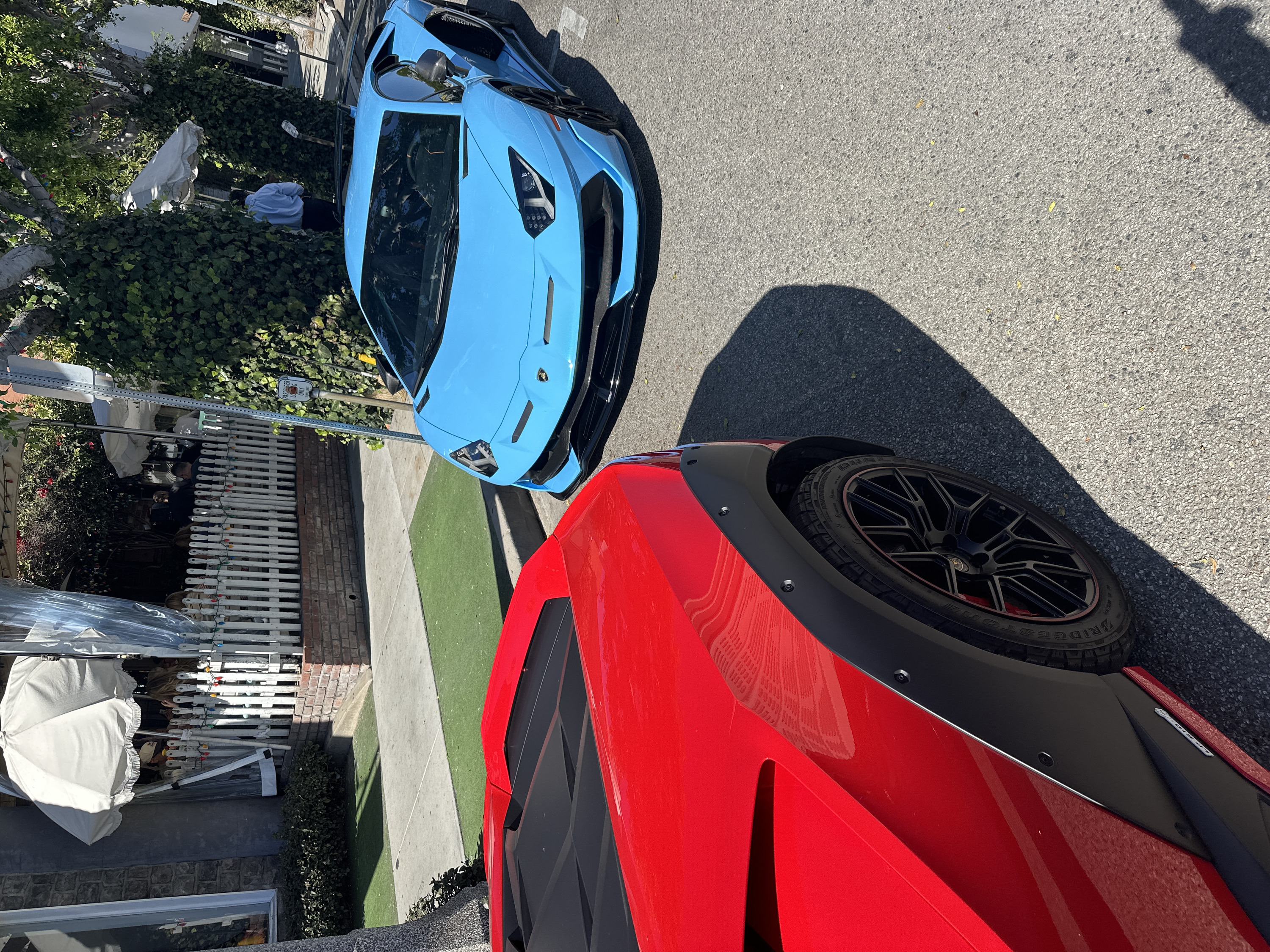 This image, rotated 90 degrees clockwise, captures a street scene featuring two high-performance sports cars parked on a sunny day.
In the foreground, taking up the lower right portion of the rotated image, is the rear section of a bright red sports car. It appears to be a Lamborghini, characterized by its aggressive styling. The car features a prominent black, wide-body fender flare, secured with visible bolts or rivets, and a multi-spoke black rim with a subtle red accent line on the tire's edge. The tire's sidewall clearly shows the brand "BRIDGESTONE." Reflections of the surroundings, possibly trees or buildings, are visible on its glossy red paintwork. A black louvered or mesh-like design is seen on the car's side, behind the wheel arch.
Slightly behind and to the left of the red car, another luxury vehicle, a bright light blue (cyan) Lamborghini, is prominently displayed. This car's front is facing the viewer, showcasing its distinct aerodynamic design, sharp LED headlights, and the Lamborghini bull emblem on its hood. The front bumper and splitter are black, providing a stark contrast to the blue body. The car's windshield and roof are also visible, reflecting the bright sky.
The cars are parked on an asphalt road. Adjacent to the road is a concrete sidewalk, followed by a strip of neatly maintained, bright green turf or grass. Beyond this green strip, dense green foliage, likely ivy, covers walls and fences. In the background, partially obscured by the foliage, is what appears to be an outdoor area belonging to a building. This area features a white picket fence, adorned with string lights, and sections of brickwork. Several white patio umbrellas are visible, some standing upright, others possibly folded or covered, along with clear plastic sheeting or tarps, suggesting an outdoor dining or lounge setup. A few individuals are faintly visible in the background, one person wearing a light blue shirt and dark pants, appearing to be standing near the fence.
The lighting in the image is bright and clear, indicating a sunny day. Strong, elongated shadows are cast by the red car and other objects on the asphalt, suggesting it is either mid-morning or afternoon. The overall impression is one of luxury and an urban or suburban setting.