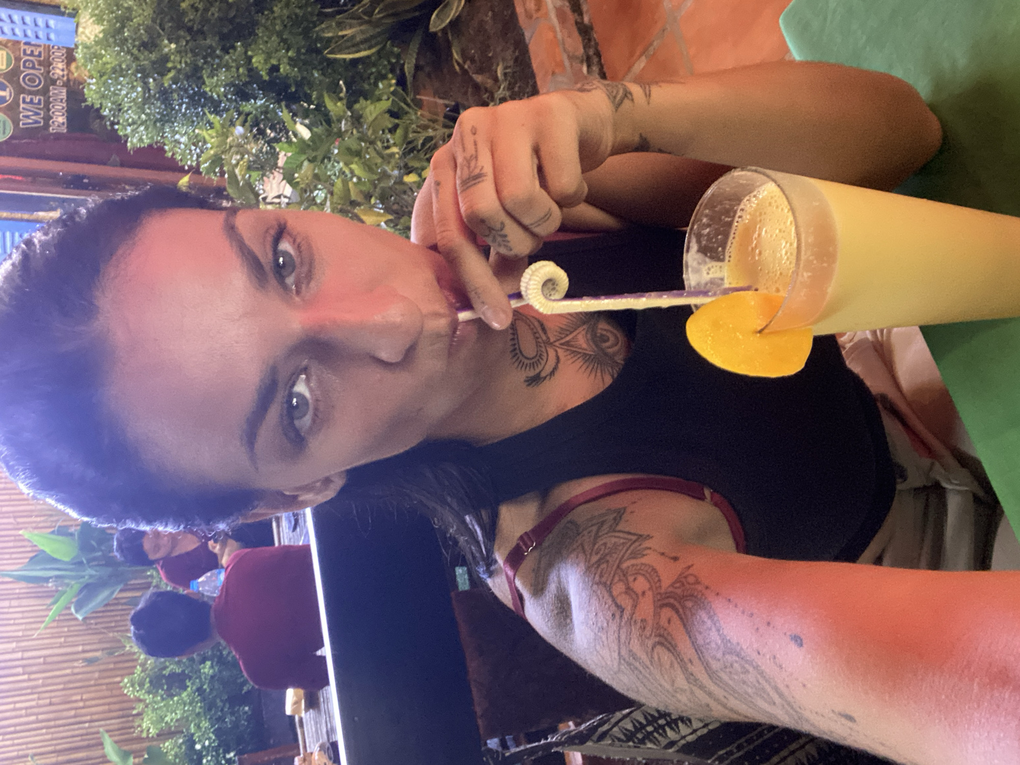 A woman with dark hair, tattoos on her arms and chest, and light-colored eyes is taking a selfie while holding a tall glass of an orange beverage with a slice of orange on the rim and a purple straw with a swirled end. She is wearing a black tank top. Behind her, to the left, two individuals are seated at a table with a bamboo wall in the background. The person in the foreground to the left is wearing a maroon shirt, and the person behind them is wearing a maroon shirt as well. To the right of the woman, there is greenery and a brick wall. The beverage is on a green surface. The scene appears to be an outdoor dining area, possibly a restaurant or cafe, as suggested by the presence of tables and seating, and the mention of Phu Thuy Ward, Vietnam, indicates an urban or semi-urban setting. The lighting suggests it is evening or late afternoon.