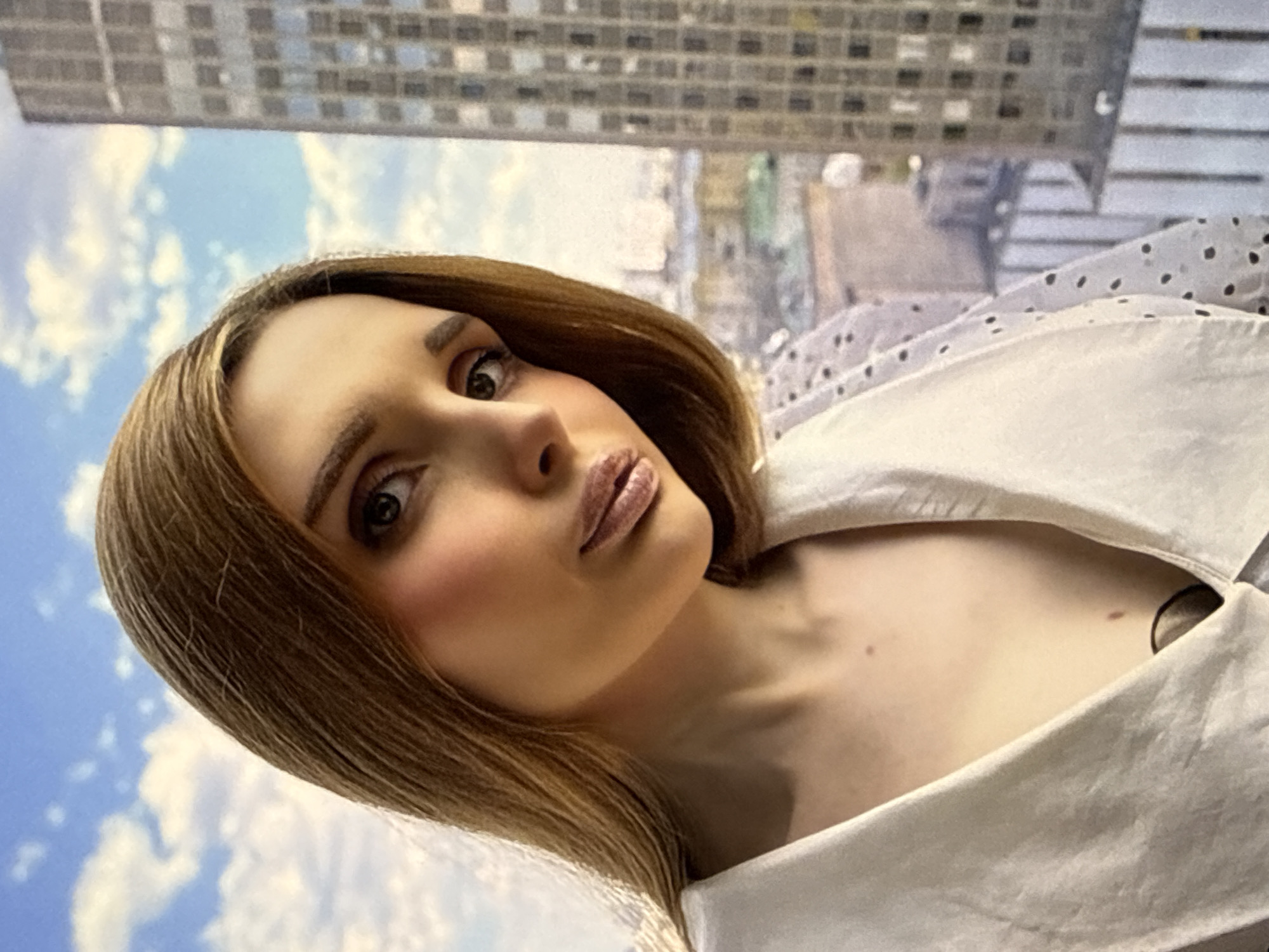 A young woman with straight, light brown hair is posed in a close-up, upper body shot, facing towards the left side of the frame with her gaze directed forward. She is wearing a white blouse with small, dark polka dots on the sleeves and a deep V-neckline. Her makeup includes subtle pink blush on her cheeks and lips, and her eyebrows are well-defined. The background features a bright blue sky with scattered white clouds, suggesting an outdoor or high-rise setting. To the right of the woman, a tall, modern building with a grid-like facade dominates the background, indicating an urban environment. The woman's left shoulder and part of her chest are visible, with two small moles visible on her skin.