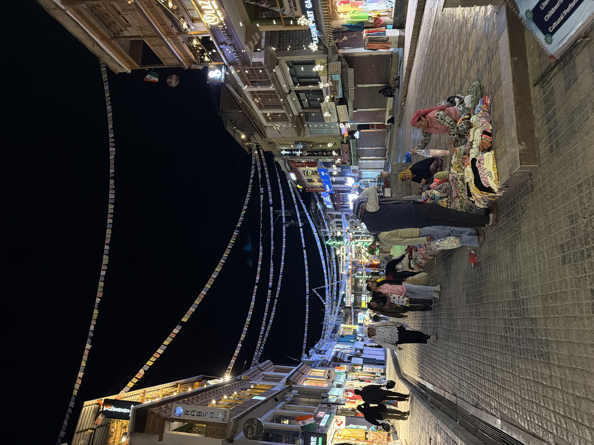 The image depicts a nighttime scene on a cobblestone street in Leh, India. The street is lined with multi-story buildings adorned with decorative lights and banners. Prayer flags are strung across the street at various heights, creating a festive atmosphere. Several individuals are present on the street. In the foreground, two women are sitting on the ground next to piles of colorful knitted goods, seemingly engaged in selling their wares. One woman, wearing a pink headscarf and a patterned jacket, is holding up a bag of items. A man, partially visible on the left, appears to be a customer or passerby. Further down the street, a group of people are walking, some carrying bags. Shopfronts with various signs, including "RABSAL CAFE", "BIRKANT QUEEN", and "KASHMIR ART GALI", are visible along the buildings. The overall impression is that of a bustling marketplace or a main street in Leh at night, with shops and vendors open for business.