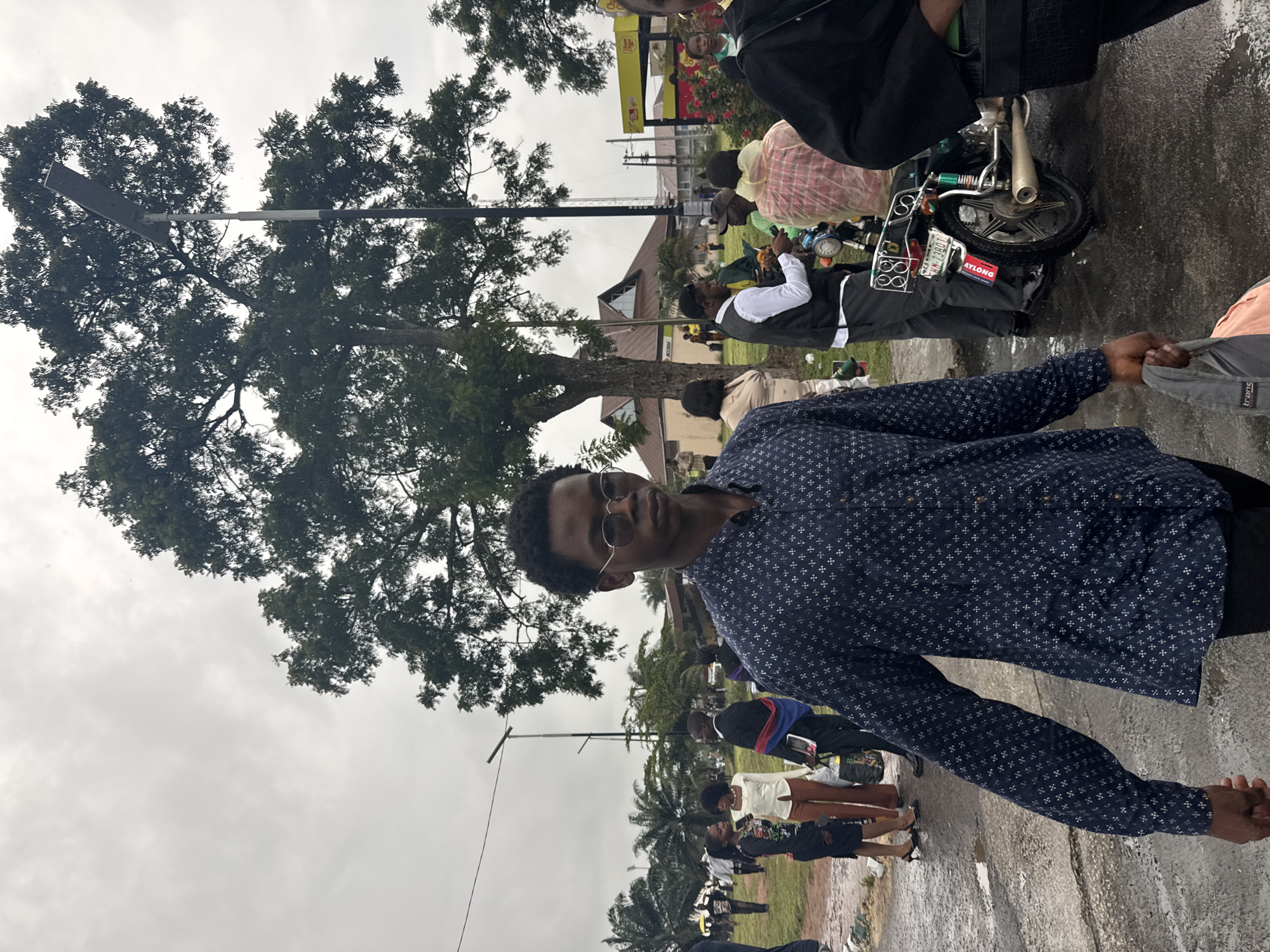 This image depicts an outdoor scene in Ihiagwa, Nigeria, under an overcast sky. The ground is dark and wet, indicating recent precipitation.
In the immediate foreground, a young Black male walks towards the viewer's left. He wears round, dark-tinted sunglasses, a long-sleeved dark blue button-up shirt with a white geometric pattern, and dark trousers. He carries a gray and peach-colored fabric bag in his right hand. To his right, the upper arm and shoulder of another individual, clad in dark clothing, are partially visible.
The midground features numerous individuals and structures. To the left of the foreground subject, a small group of people is present. Two women stand conversing; one wears a floral long-sleeved top and dark skirt with dark high heels, and the other wears a cream-colored long-sleeved top and light brown trousers. Near them, a man in a dark suit with a red and blue striped fabric draped over his left shoulder holds a dark object. A tall streetlight with a rectangular solar panel-like fixture is visible behind this group. Further back on the left, several palm trees are distinguishable.
In the midground-right, a group of individuals gathers around a dark-colored motorcycle. One man wears a white long-sleeved shirt and a dark vest. Another person is covered by a translucent red and white checkered garment, possibly a rain poncho. The rear of the motorcycle is visible, featuring a license plate with "IMO H-138DT" and a vertical "NYONG" sticker on its side. It has a chrome exhaust pipe and a metal rear rack.
The background is characterized by dense green trees. A multi-gabled building with a brown roof and an "ACCESS" sign is visible in the central-left background. To the right, a yellow structure with "Chicken George" text is partially visible, indicating a commercial establishment. Additional distant buildings and structures are discernible beneath the light gray, overcast sky.
The ground surface consists of dark, wet asphalt or concrete, showing cracks and containing visible puddles.