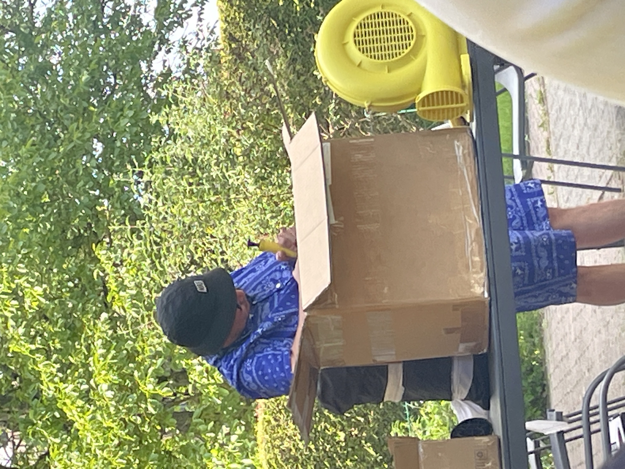 A man in a black baseball cap and blue paisley shirt is inserting a yellow tool into a cardboard box. The box is positioned on a metal frame structure, adjacent to a yellow air blower. A second box is partially visible below. A green foliage background is present, suggesting an outdoor setting. The location is Wienhausen, Germany.