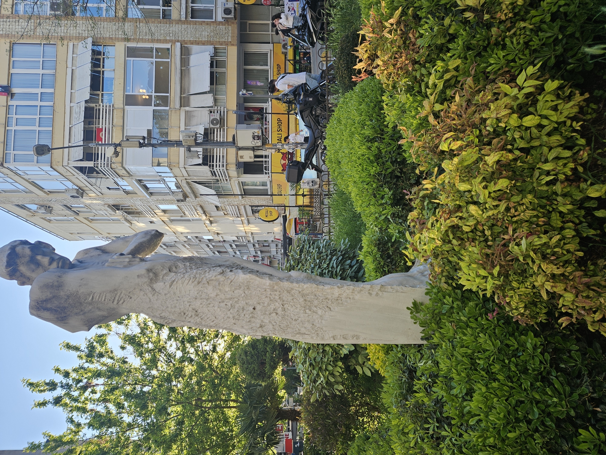 The image shows a statue, seemingly of a reclining figure, situated in a park or green space within the urban environment of Kadıköy, Turkey. Behind the statue, there is a multi-story building with numerous windows and air conditioning units. A few people are present in the background, some possibly riding motorbikes. Businesses are identifiable in the background with yellow signage. The foreground is filled with green shrubbery. The sky is visible behind the trees.