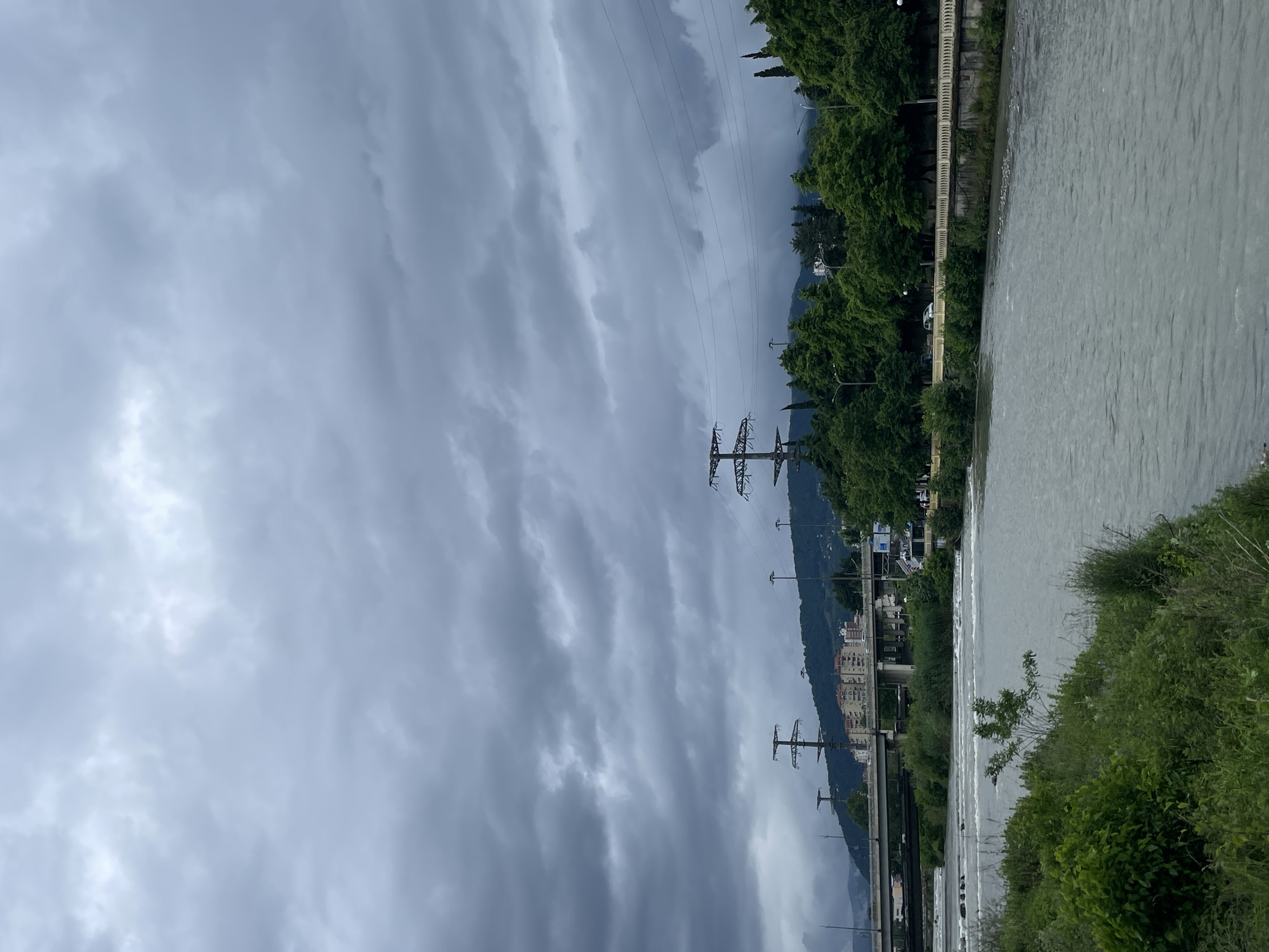 The image shows a river landscape with an overcast sky, likely in Sochi, Russia. The river's water appears turbid and flows from right to left. Along the right bank of the river, there's dense green vegetation. A concrete structure, possibly a bridge or a walkway, is situated alongside the river.
In the background, partially obscured by trees, there are buildings suggesting an urban environment. Power lines and pylons stretch across the frame, adding a linear element to the landscape. Mountains are visible in the distance behind the buildings. The heavy, layered clouds fill most of the sky, indicating possible inclement weather.
