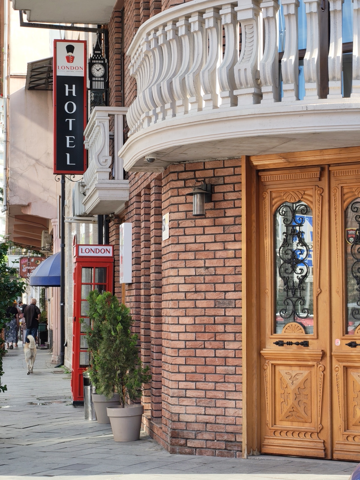 The image depicts the exterior of a hotel in Batumi, Georgia, styled with a London theme. A prominent vertical sign with "HOTEL LONDON" in white lettering on a black background, topped with an illustration of a British guard and a Big Ben clock tower, hangs from the building. Next to the sign is a bright red, traditional British telephone booth. The building features a red brick facade and a decorative white balustrade above a large, ornate wooden double door with carved panels and decorative metal grilles on the glass inserts. A vintage-style outdoor light fixture is mounted on the brick wall. The sidewalk in front of the hotel is paved with grey tiles. Two potted evergreen trees stand near the entrance. In the background, a few pedestrians are walking along the sidewalk, and a dog is seen walking away from the camera. The overall scene conveys a blend of European architectural influences with a distinct London theme.