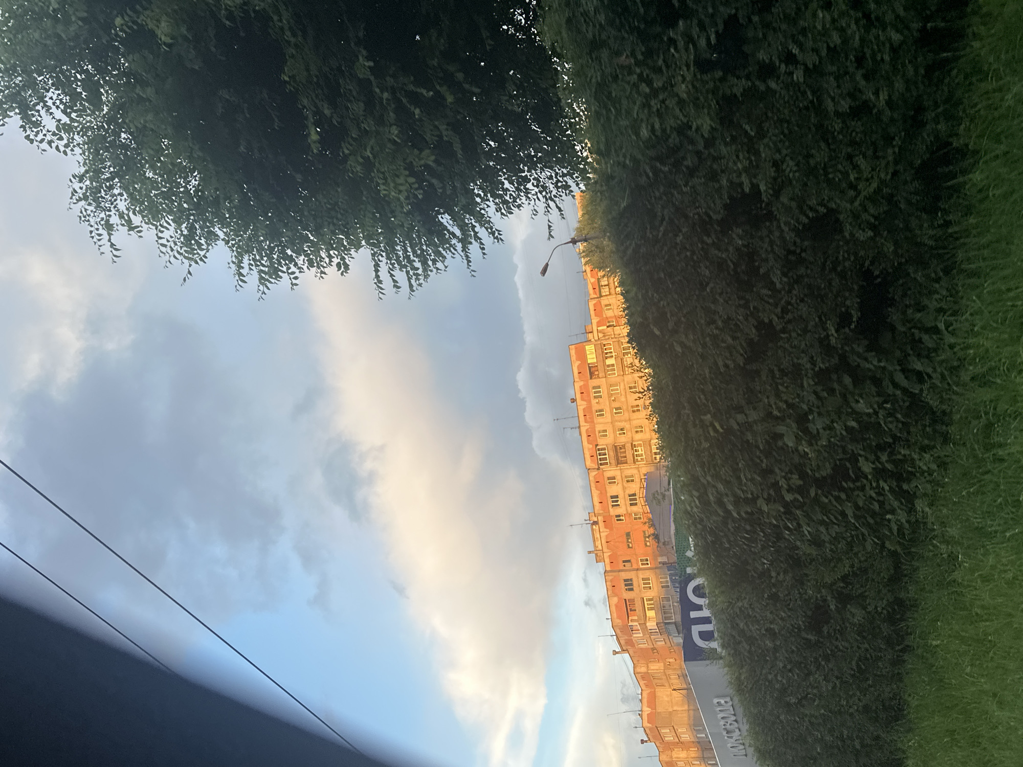 This is a rotated photograph taken from a low angle, likely from within a vehicle, looking up and outwards. The scene is dominated by a building with many windows, appearing to be an apartment block, rendered in shades of orange and yellow. The building is situated along a street lined with dense green bushes and grass. Above the building, the sky is a mix of blue and grey clouds, with some patches illuminated by the warm glow of sunset. The time of day is late afternoon or early evening, as suggested by the golden light on the building and the clouds.
In the foreground, obscuring the lower left of the image, is a dark, out-of-focus area that appears to be the interior of a car or a similar enclosed space. Two dark, diagonal lines, possibly power lines or cables, stretch across the sky. A street lamp is visible above the building, suggesting an urban or suburban setting. The overall mood is somewhat tranquil, with the soft light and the contrast between the natural elements (trees, grass, sky) and the man-made structure.
There is visible text on the side of the building, appearing to be in Cyrillic script. Part of the text reads "ПОЧТА РОССИИ" which translates to "Russian Post". The image was taken in Magnitogorsk, Russia.