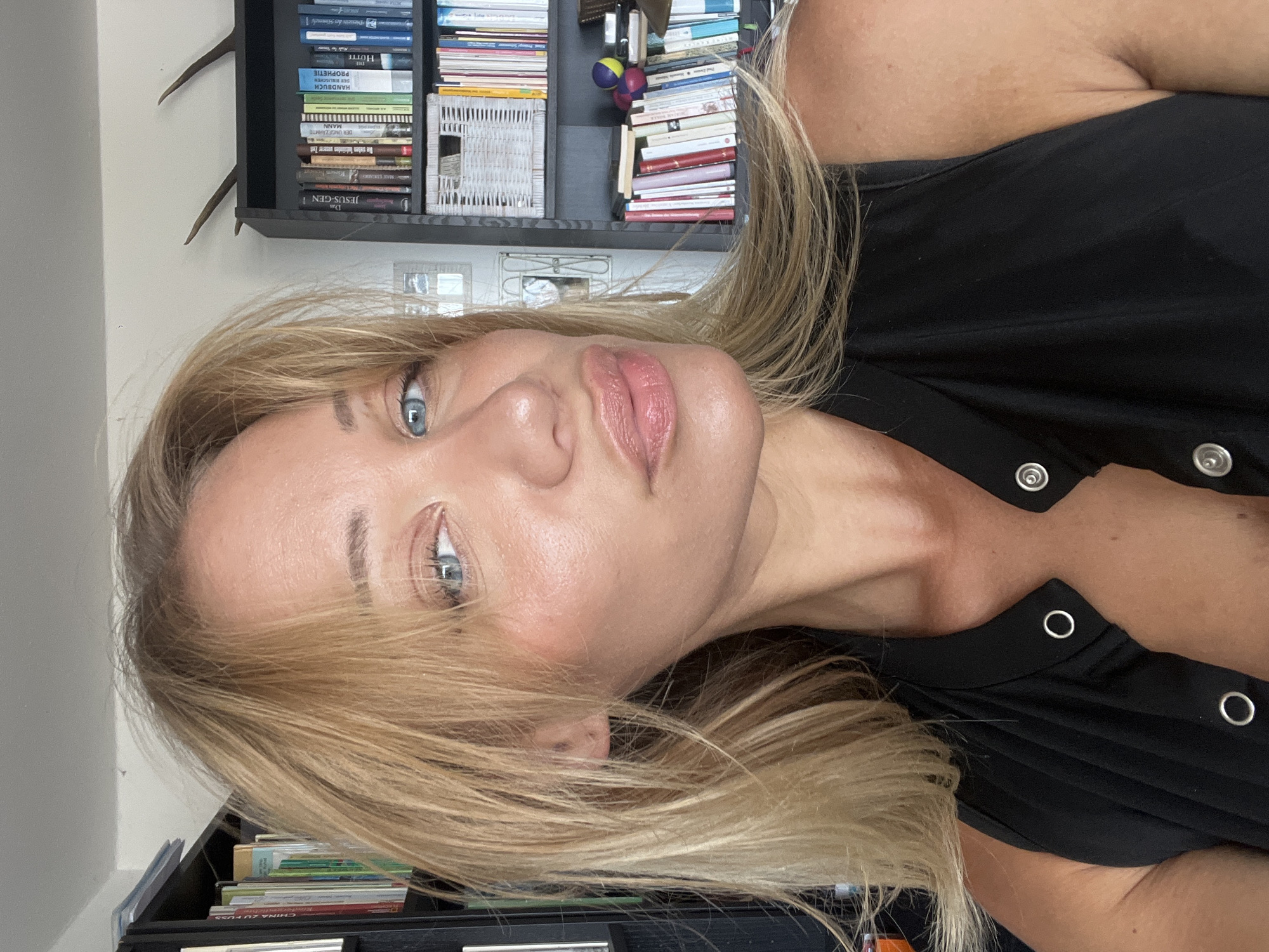 The image is a selfie of a blonde woman with blue eyes, looking directly at the camera. She is wearing a black sleeveless top with silver snaps. The background shows bookshelves filled with books, a white woven basket, and a pair of deer antlers mounted on the wall. The setting appears to be indoors, possibly a home or office. The woman's makeup is noticeable, with blush and lip gloss. The lighting suggests it is daytime. There is no information to indicate the location in Mühldorf a. Inn, Germany.