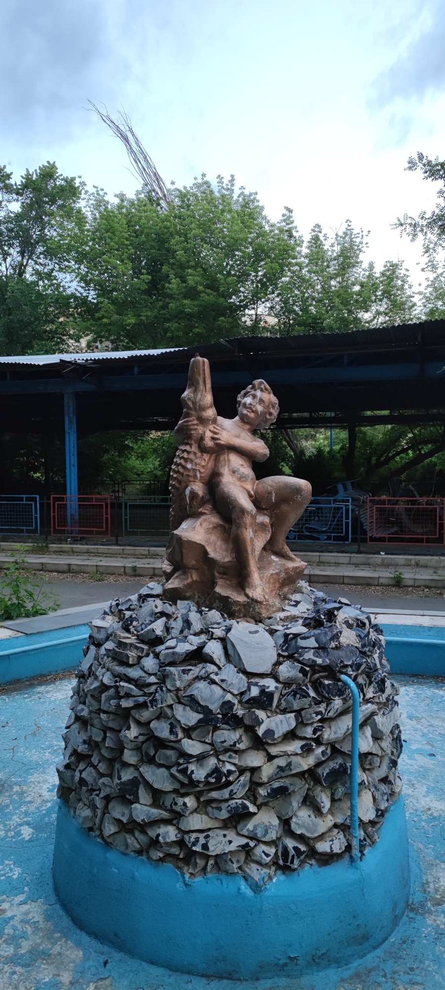 The image depicts a bronze statue of a cherubic child embracing a crocodile, situated atop a fountain base constructed from stacked stones. The fountain is painted a vibrant blue and appears to be outdoors, with trees and a covered structure visible in the background. A blue pipe extends from the base of the fountain. The statue itself is somewhat weathered, with white residue present on the child's face and chest. The surrounding area shows signs of wear, with chipped paint on the fountain and a paved walkway. The sky is overcast with a few thin branches extending from the top of the frame, suggesting a slightly neglected or aged park setting. The statue appears to be a decorative element within the city of Yerevan, Armenia.
