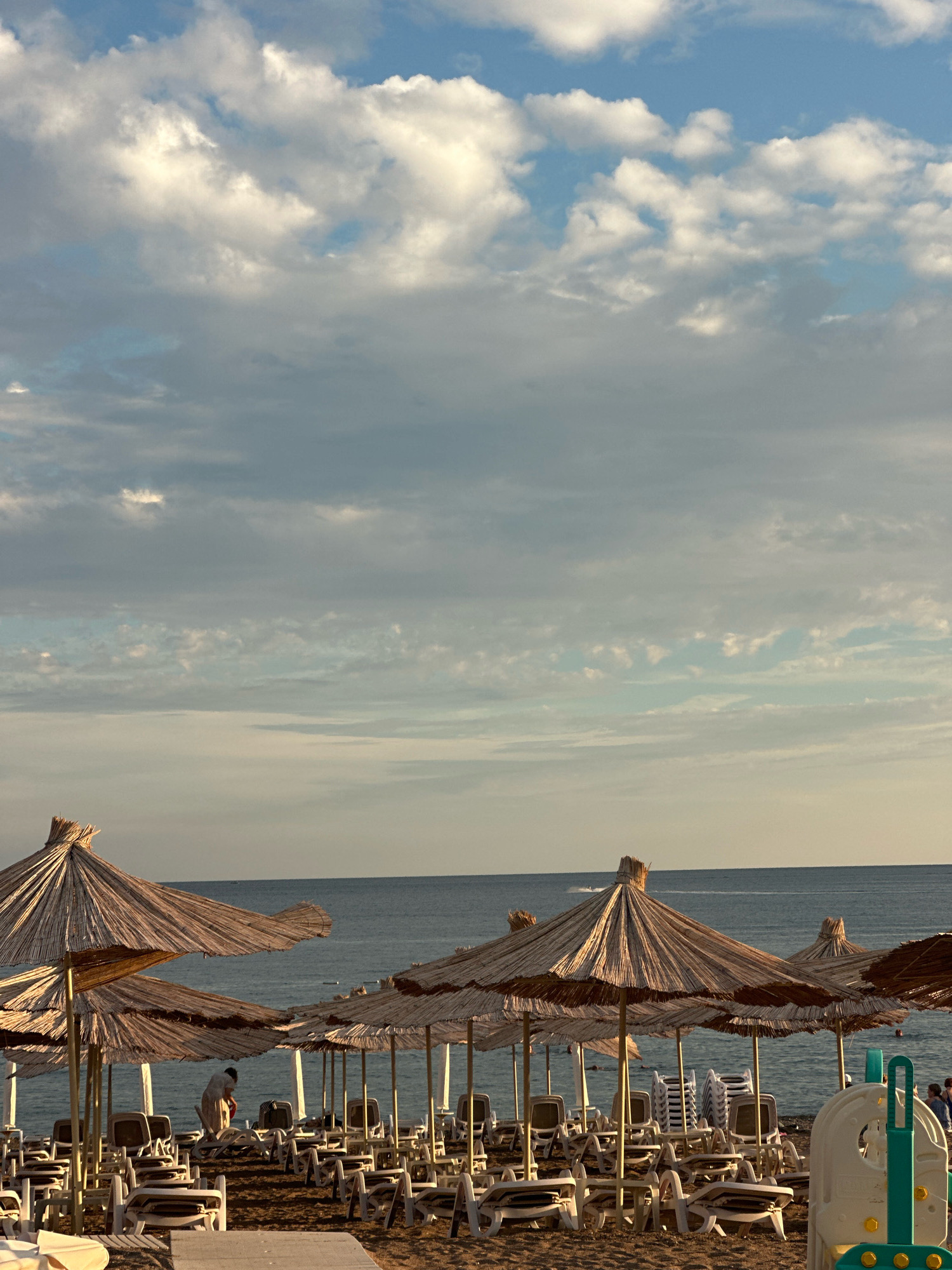 A beachfront scene in Boreti, Montenegro, shows numerous thatched-roof umbrellas and stacked lounge chairs on a sandy beach. The calm sea stretches to the horizon, under a sky filled with white and gray clouds. Several people are visible on the beach and in the water, and a watercraft is leaving a wake on the sea. The lighting suggests it is late afternoon or early evening.