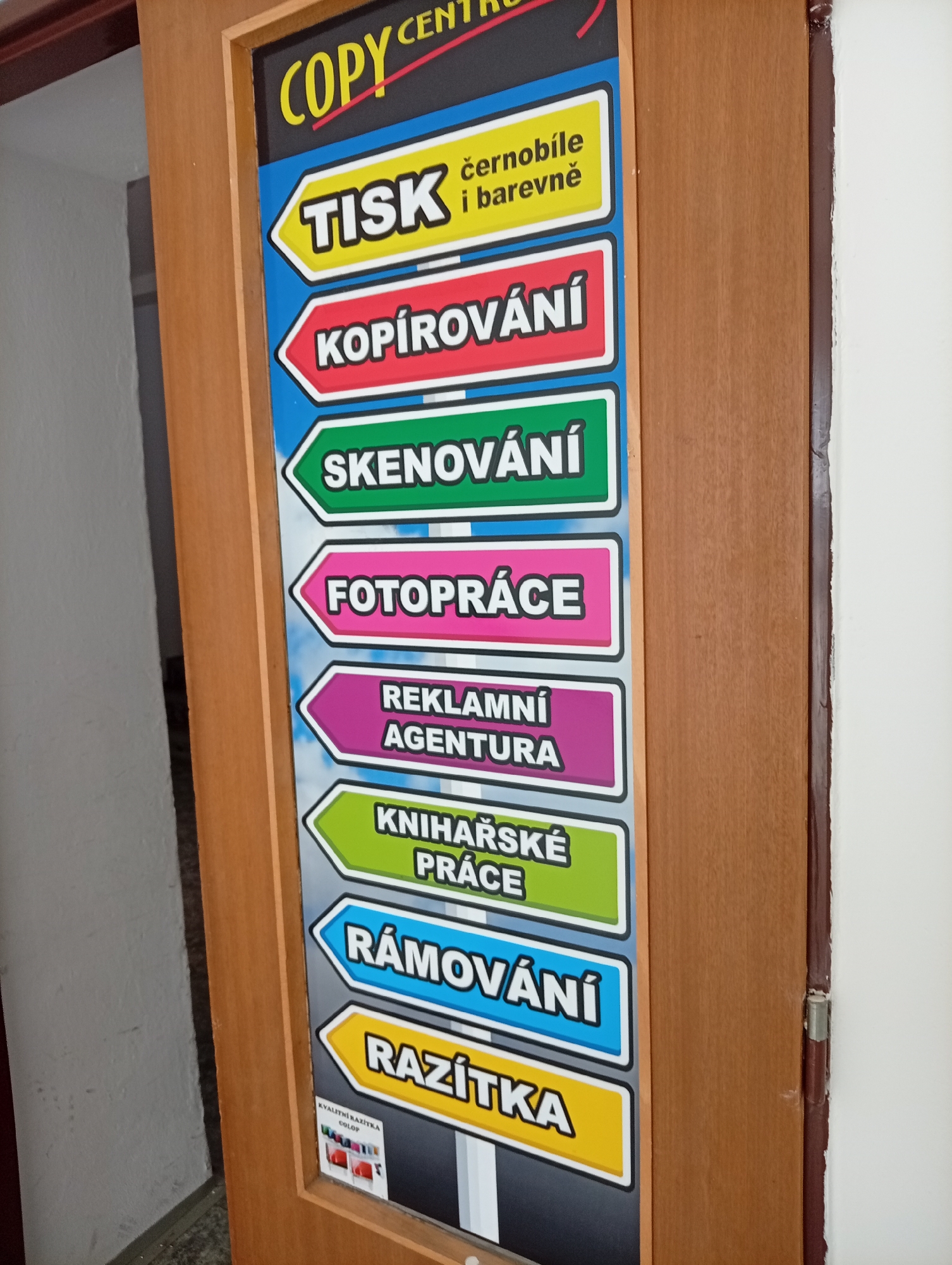 The image shows a sign board on a wooden door. The sign board lists various services offered by a copy center. The services include "TISK" (printing) in black and white and color, "KOPIROVÁNÍ" (copying), "SKENOVÁNÍ" (scanning), "FOTOPRÁCE" (photo work), "REKLAMNÍ AGENTURA" (advertising agency), "KNIHAŘSKÉ PRÁCE" (bookbinding work), "RÁMOVÁNÍ" (framing), and "RAZÍTKA" (stamps). The sign board is colorful, with each service listed on a different colored background and shaped like a directional arrow. At the bottom of the sign board, there is a small image of colorful stamps. The wooden door is light brown and appears to be ajar, revealing a glimpse of a white wall on the left side and a white wall on the right side. The overall setting suggests an interior of a business establishment in the city of Tišnov, Czechia.