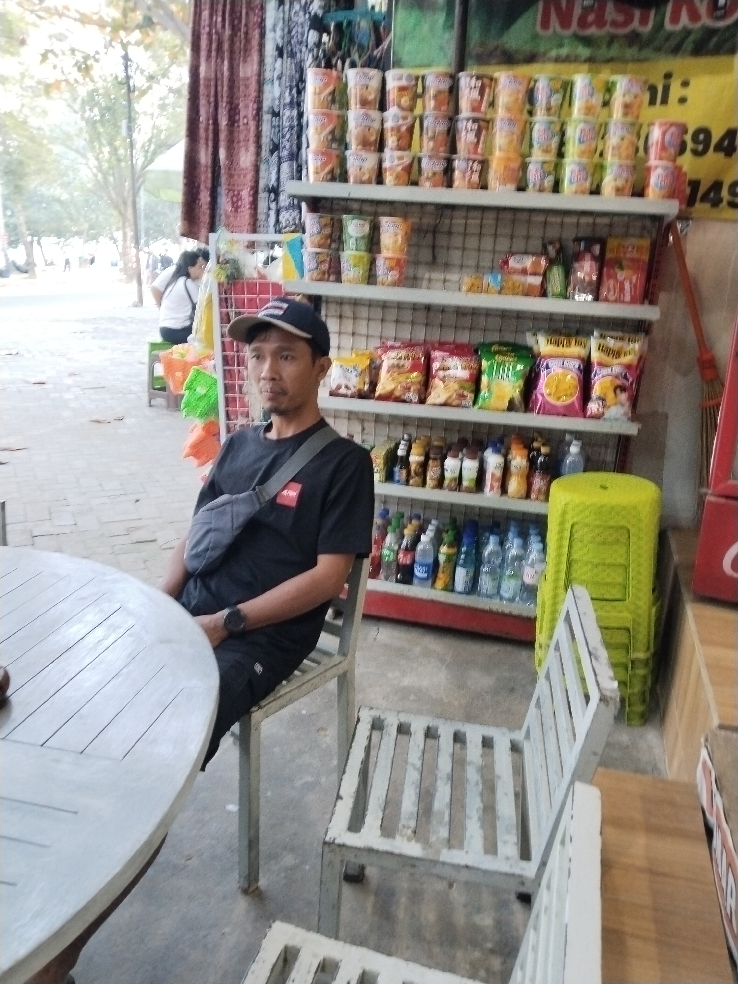 In the city of Prigi, Indonesia, a man wearing a black t-shirt and a black baseball cap sits on a metal chair in front of a round wooden table. He has a grey crossbody bag slung over his shoulder. Behind him, a shelf displays various snacks and bottled drinks, with a bright green plastic stool stacked on top of another one. In the background, people are visible on a street with trees and a building. The scene appears to be an outdoor food stall or a small shop.