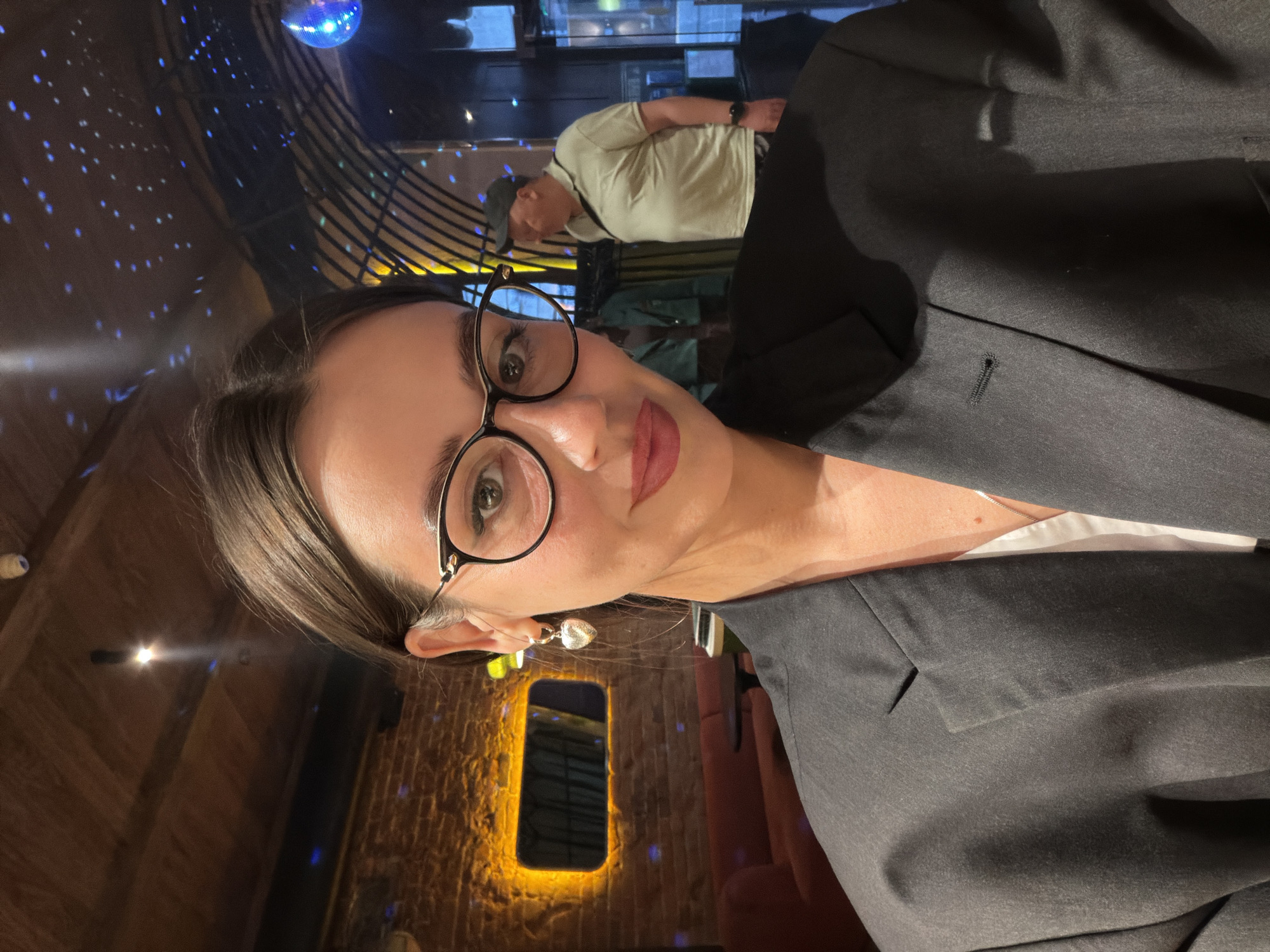 This is a close-up, rotated selfie of a woman wearing glasses and a dark blazer. She has dark hair and appears to be wearing lipstick. A heart-shaped earring is visible on her left ear. The background suggests an indoor setting, possibly a bar or club, with a disco ball and blue lights casting a starry effect. In the blurred background, another person is visible wearing a light-colored t-shirt and a cap. The overall mood seems casual and perhaps social. The image is taken from a low angle, emphasizing the subject's face. The location context is Krasnoyarsk, Russia. No text is visible in the image.