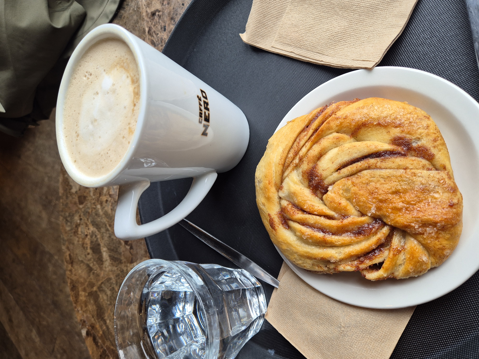 The image displays an overhead view of a table setting on a dark grey, textured tray. A white ceramic mug with the black "CAFFÈ NERO" logo contains a light brown beverage topped with white foam, positioned on the right side of the tray. To its left, a clear, faceted glass holds clear water. A slender, silver-colored utensil is partially visible between the glass and the mug. A round white plate at the bottom of the tray holds a golden-brown, twisted pastry, exhibiting a visible cinnamon swirl and sprinkled sugar crystals. Two folded, light brown paper napkins are situated on the tray: one to the left of the plate and another to the right of the mug. The tray rests on a mottled brown and beige surface, resembling marble. A dark olive-green fabric object is visible in the upper right background. The location is identified as a Caffè Nero establishment, a chain that operates within the West Suffolk district of the United Kingdom.