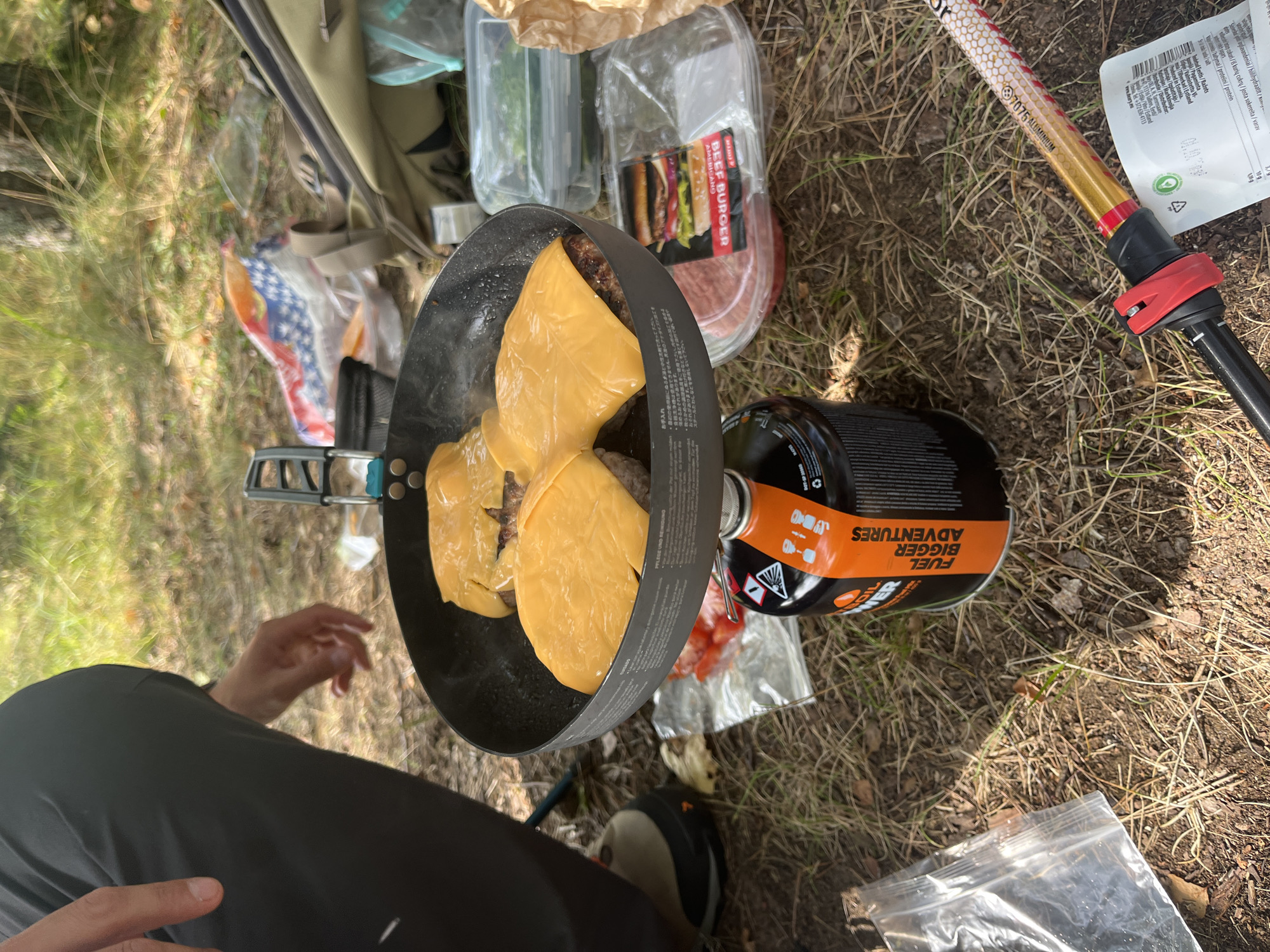 In Koitjärve, Estonia, a person is preparing cheeseburgers on a portable camping stove. The scene is outdoors, with dry grass and pine needles covering the ground. A black frying pan, containing three beef patties topped with melted yellow cheese, is placed on the stove. The stove is a black and orange canister labeled "FUEL BIGGER ADVENTURES". Nearby, there are plastic containers holding what appears to be chopped tomatoes and greens, as well as a package of raw beef patties and a bag of what looks like burger buns. A person's dark gray pants and a hiking boot are visible on the left side of the frame. A trekking pole lies diagonally across the lower right corner of the image. The overall atmosphere suggests a camping or outdoor meal preparation in a natural setting.