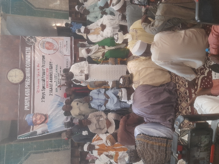 The image shows a gathering of men in a room with a carpet floor, possibly in a mosque or a community hall. The men are dressed in traditional Nigerian clothing, predominantly white robes and caps, with some wearing colorful scarves or headwear. Many are seated on the floor, with several standing around them. A man in a white robe and cap is standing and speaking to the group. Behind him, a large banner hangs on the wall, displaying the text "Prayer and Preaching Committee." There is a clock above the banner. The image is taken from a low angle, with a phone or a camera positioned in the foreground. The coordinates (latitude: 12.167, longitude: 6.659) indicate that this scene is likely located in a building within the city of Gusau, Nigeria.