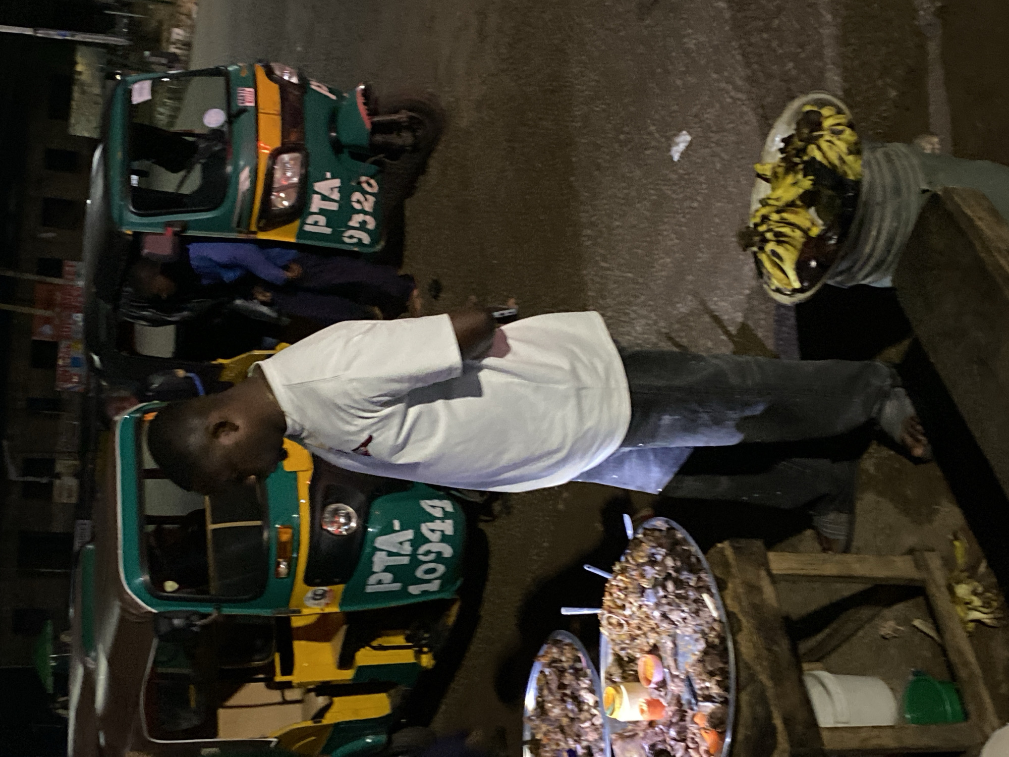 A nighttime street scene in Bukuru, Nigeria, features two auto-rickshaws, known locally as "Keke," parked on an asphalt road. The Keke on the left is predominantly yellow and green, with the license plate "PTA-10944" visible on its side. The Keke on the right is also green and yellow, displaying the license plate "PTA-9320."
A man, wearing a white t-shirt and dark trousers, stands between the two Keke, facing away from the viewer. He appears to be interacting with the scene or observing something. To the left of the man, a wooden table holds two large metal platters filled with food, possibly meat dishes, along with small bowls containing condiments. Below the table, a white bucket and a green bucket are visible.
To the right of the man, a large, light-colored bucket is topped with a platter of bananas and other dark, unidentified fruit. The background is dimly lit, showing the outlines of buildings and suggesting a commercial or roadside vending area. The overall atmosphere is one of a busy street market at night.