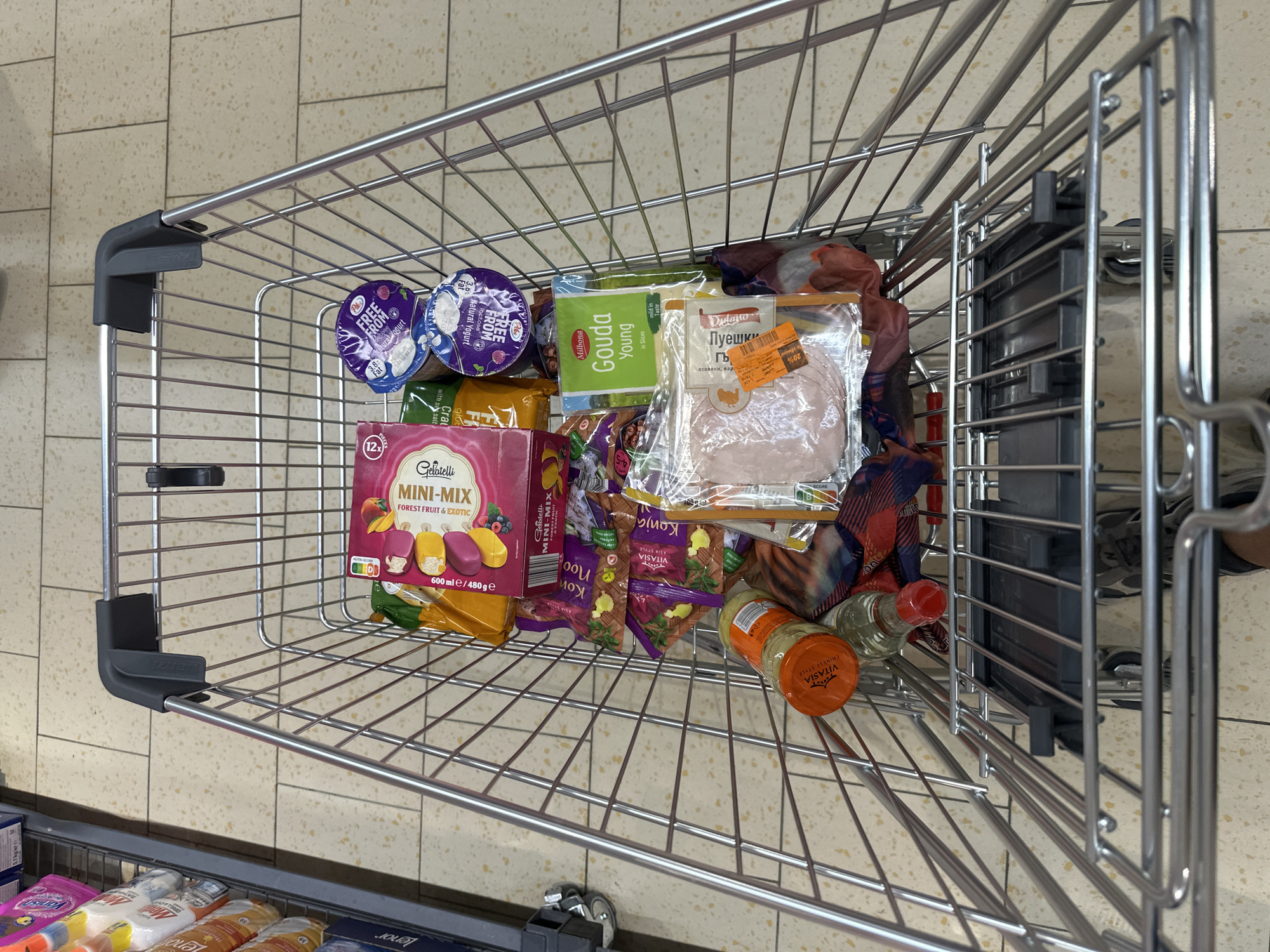 A shopping cart filled with groceries is shown from a high-angle perspective. The cart is made of silver metal bars and contains various food items. Among the visible items are: a box of "Gelatelli Mini-Mix Forest Fruit & Exotic" ice cream, two purple yogurt cups labeled "Free From Natural Yogurt", a green package of "Milbona Gouda Young" cheese slices, a package of sliced ham, and several small bags of "Vitasia Kanja Asia Style" snacks. Two bottles, one with an orange label and one with a red cap, are also in the cart. The floor is tiled with light-colored square tiles. The scene is likely in a supermarket in Sofia, Bulgaria, based on the presence of products from European brands and the overall shopping environment.