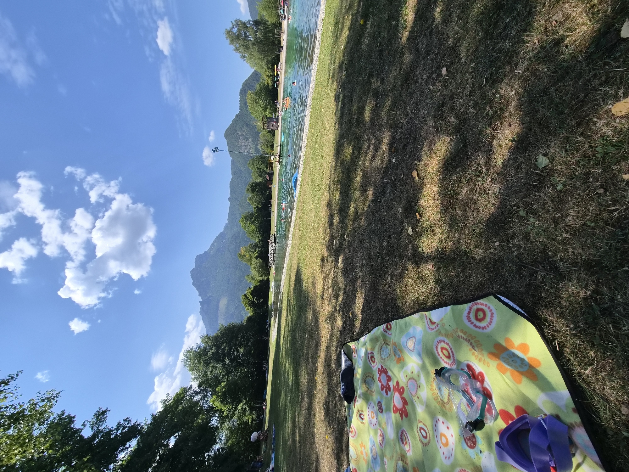 The image depicts a sunny day at a public swimming area in Landl, Austria. In the foreground, a colorful picnic blanket with a floral pattern is laid out on a grassy area. On the blanket, there are a pair of swimming goggles and a purple swim cap. Beyond the picnic blanket, a large body of water, likely a lake or a well-maintained outdoor pool, is visible. Several people are in the water, some swimming and others relaxing on inflatable devices. The water is a clear turquoise color. Surrounding the water is a manicured grassy area with trees. In the background, a mountain range dominates the horizon under a bright blue sky with scattered white clouds. The overall scene suggests a recreational setting for swimming and relaxation during pleasant weather.