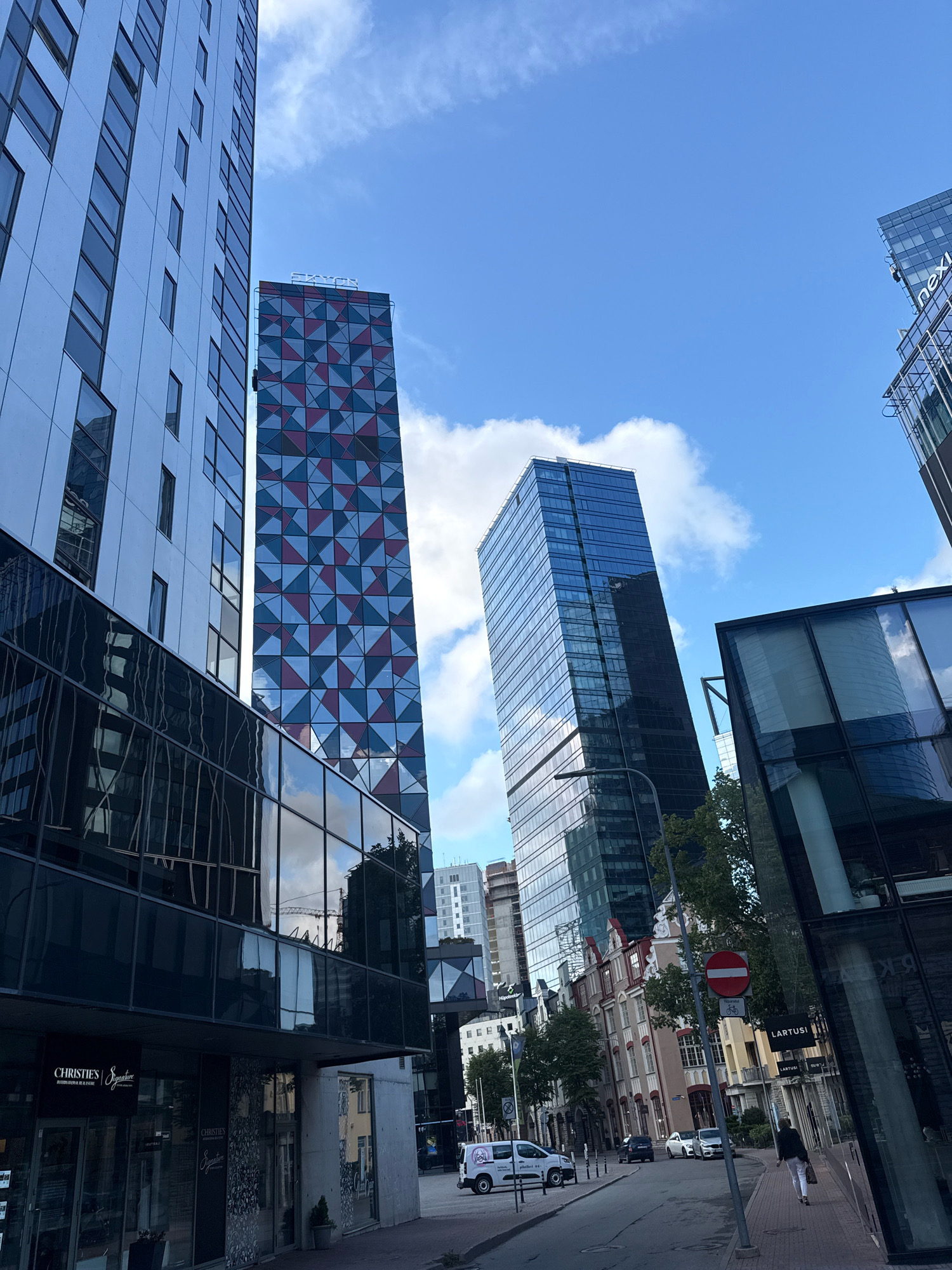 The image depicts a street scene in Tallinn, Estonia, likely in a modern business district. Tall, contemporary glass and steel buildings dominate the skyline, interspersed with a few older, more traditional European-style buildings.
In the foreground on the left, a large building with a reflective glass facade and light-colored panels is visible. Adjacent to it is a taller skyscraper with a distinctive geometric facade in shades of red, blue, and white. The top of this building has a white illuminated sign that reads "EXXON". Further to the right, a very tall, slender skyscraper with a dark blue glass exterior stands prominently.
On the ground level, several businesses are visible. One storefront has signage that reads "CHRISTIE'S Signature".
The street is paved with asphalt and lined with parked cars and a white van. A few individuals are walking on the sidewalk. A traffic sign with a red circle and a horizontal white bar is mounted on a pole, indicating no entry. Trees and foliage are present along the street, suggesting a mild climate. The sky is blue with scattered white clouds. The overall impression is one of urban development, with a mix of new and old architecture.