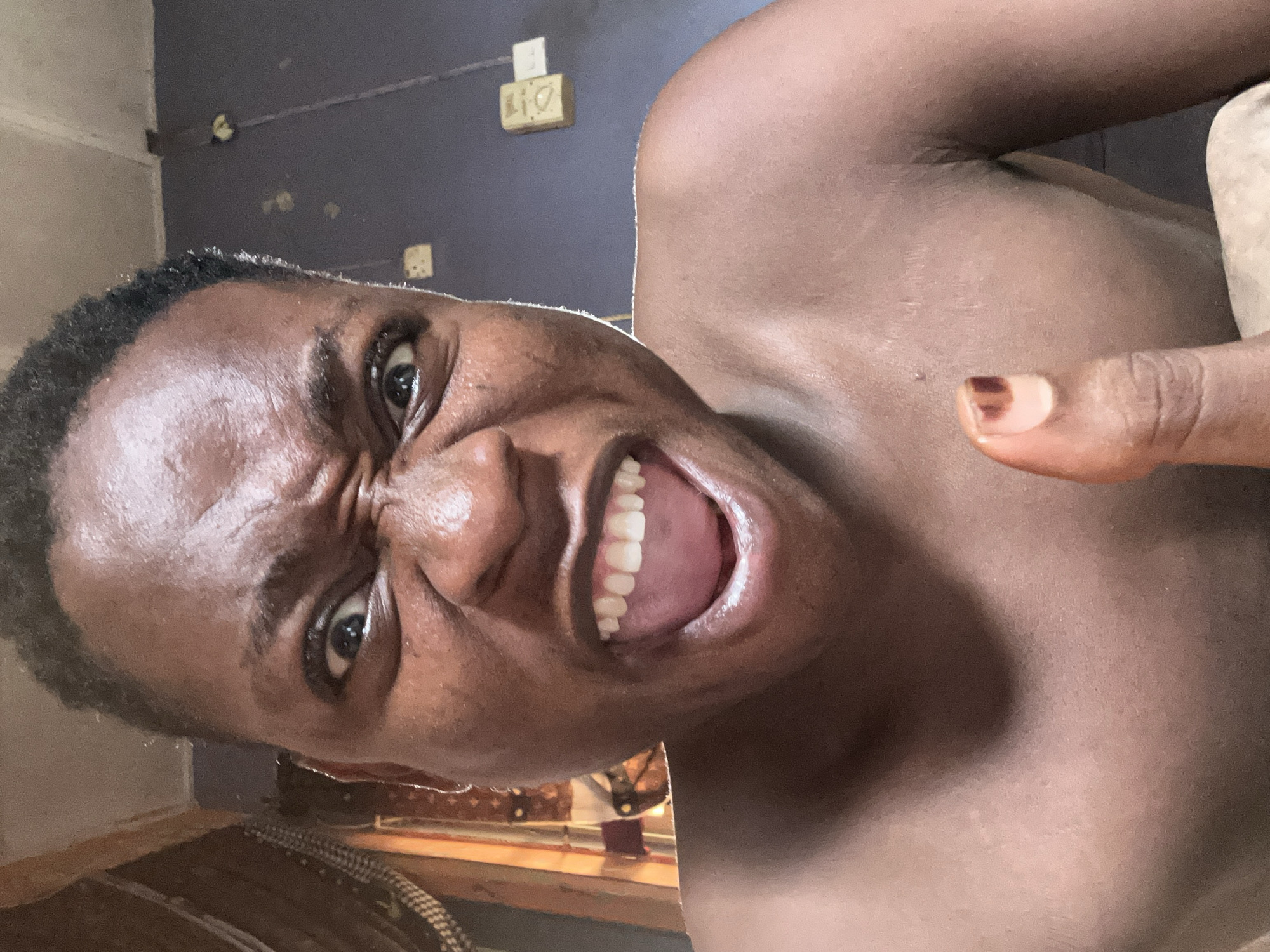 A close-up, tilted selfie shows a dark-skinned person with short, dark hair making an angry facial expression, including a wrinkled nose and bared teeth. The person's finger is visible in the top right corner of the frame. The background is a dark blue wall with an electrical outlet visible. The location is likely indoors, within the city of Ikorodu, Nigeria.