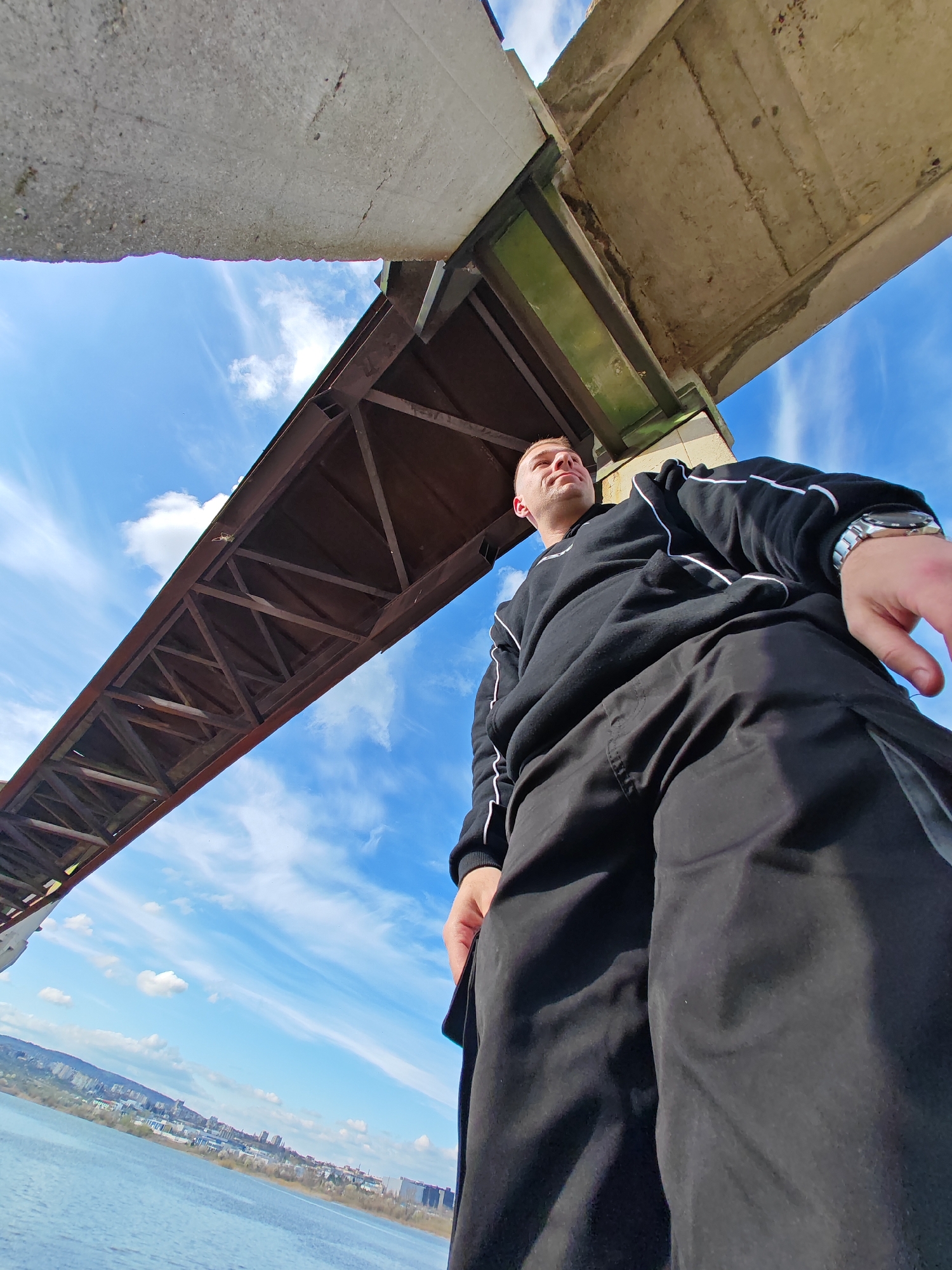 A low-angle shot features a man standing beneath a bridge. The bridge is constructed of concrete and steel, with a visible truss structure. The sky is light blue with scattered clouds. In the background, across a body of water, is a cityscape. The man is wearing a dark sweatshirt with white stripes on the sleeves and dark pants. He has a watch on his left wrist. The location is Shumen, Bulgaria.